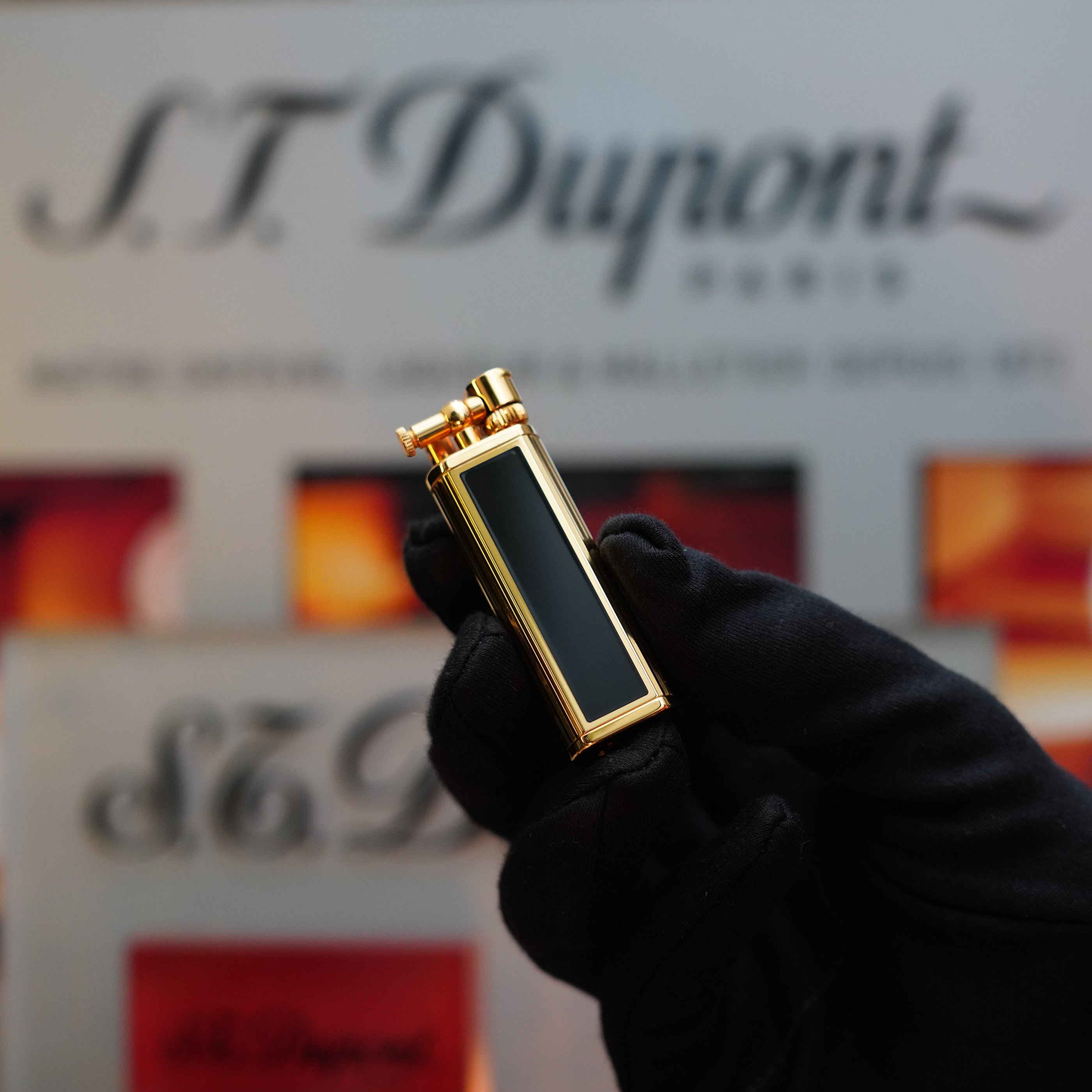 A gloved hand holding a 1980 Dunhill Unique Sylfide 24k Heavy Gold Natural Lacquer Lighter in front of a blurred S.T. Dupont Paris sign.