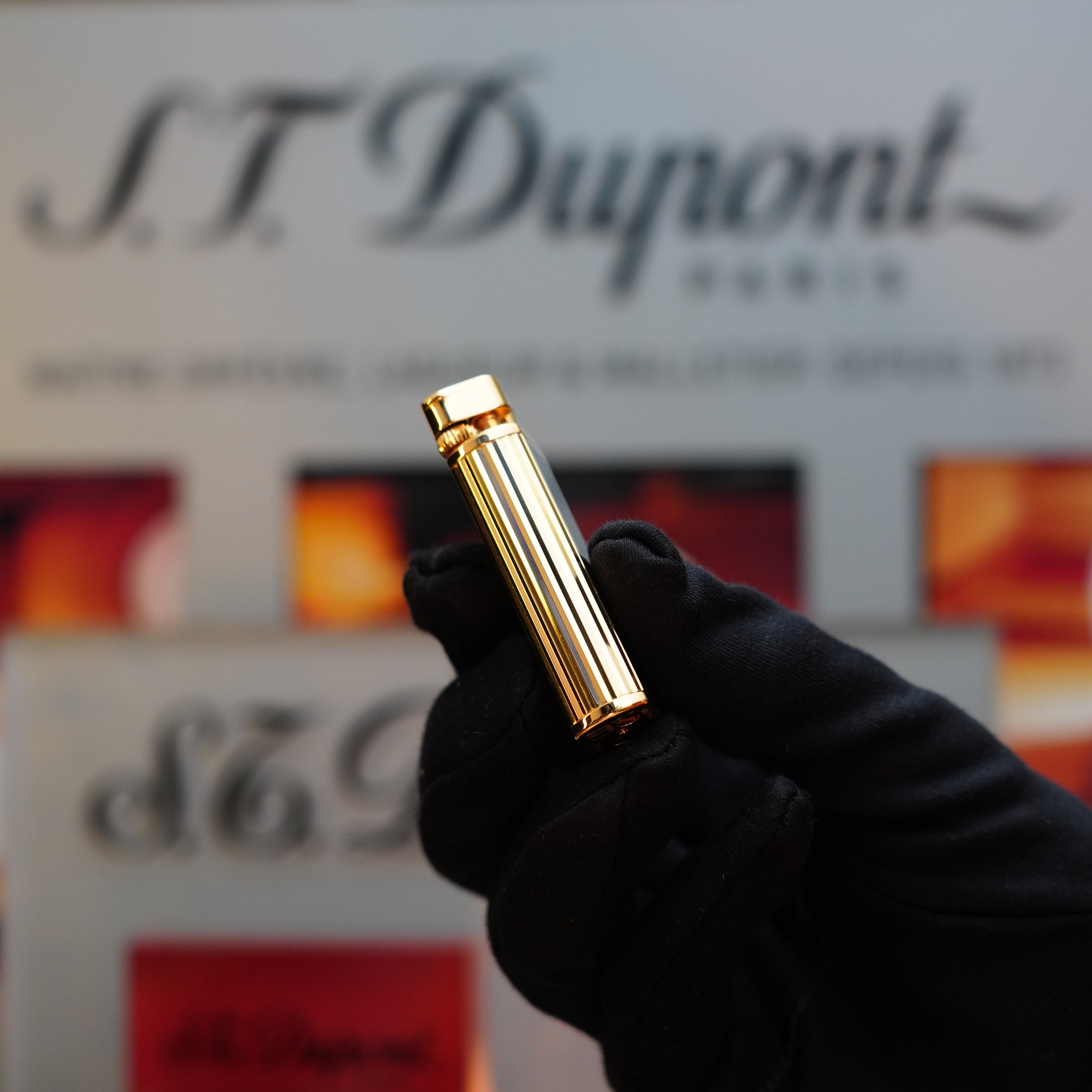 A gloved hand holds a gold cylindrical object, resembling the 1980 Dunhill Unique Sylfide 24k Heavy Gold Natural Lacquer Lighter, in front of an out-of-focus background displaying the text "Dunhill.