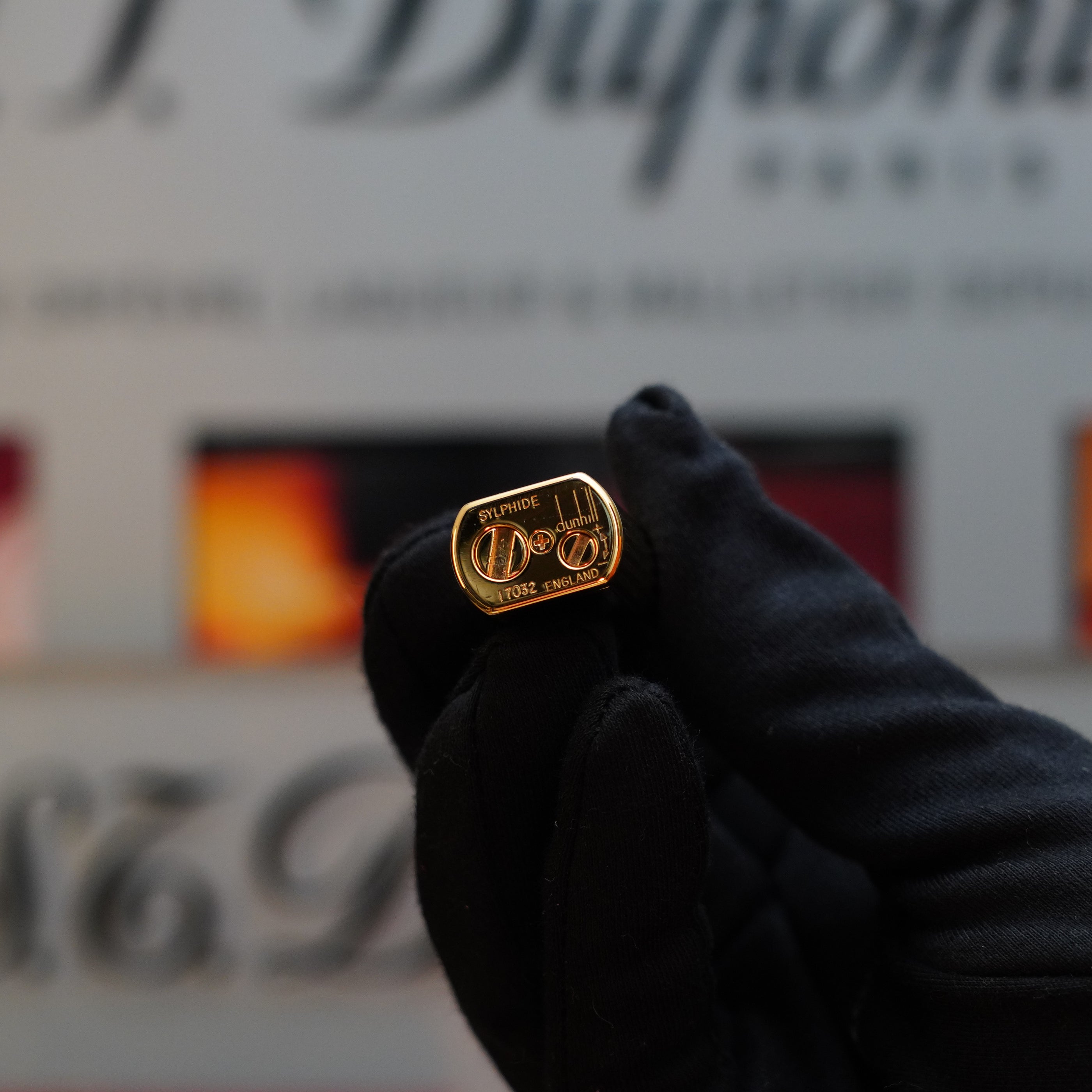 A gloved hand holds a vintage lighter, specifically the 1980 Dunhill Unique Sylfide 24k Heavy Gold Natural Lacquer Lighter by Dunhill, against a blurred background. Engraved on the lighter are the letters "D" and "C" with some additional text.