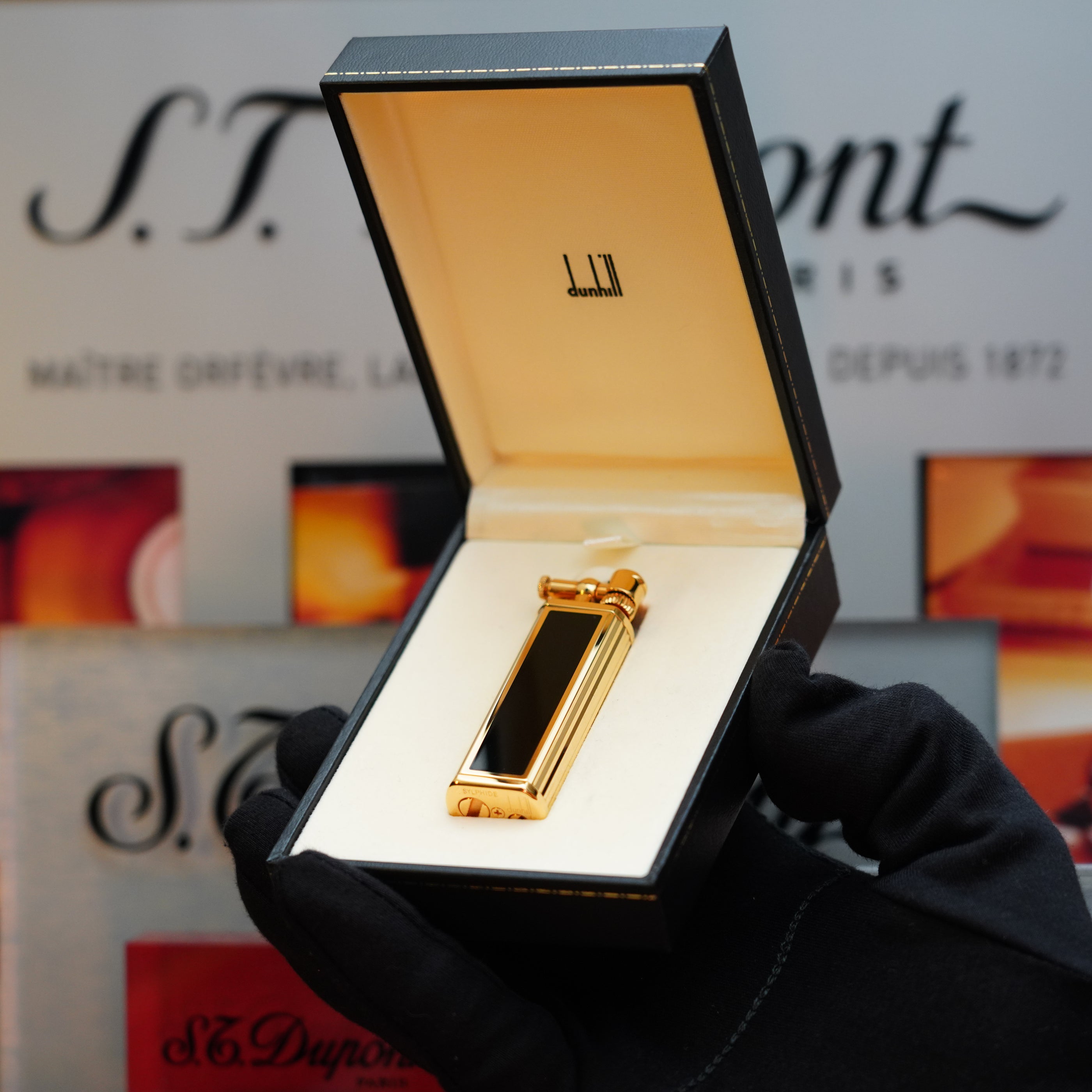 A 1980 Dunhill Unique Sylfide 24k Heavy Gold Natural Lacquer lighter with "Dunhill" branding displayed inside an open box held by a gloved hand.