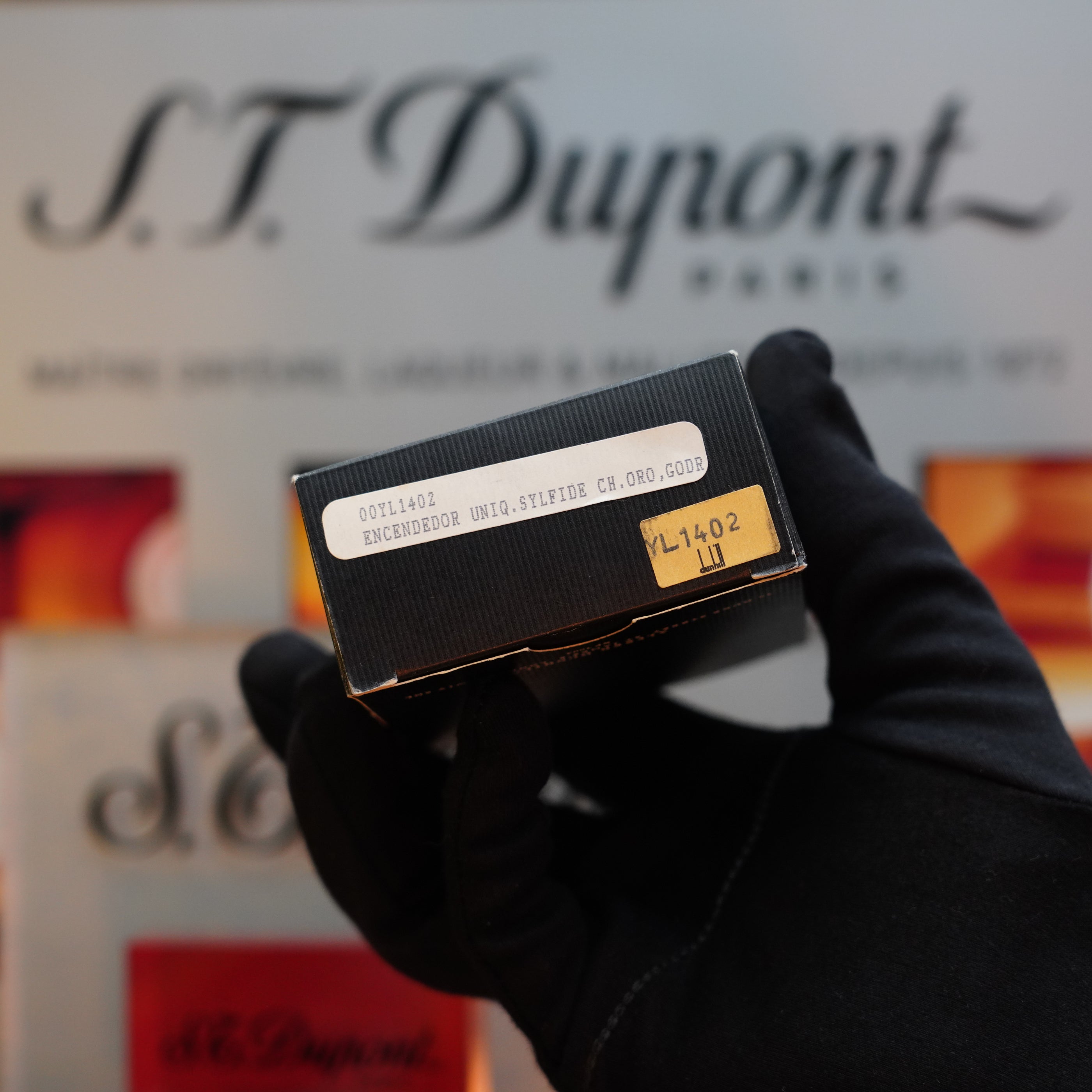 A gloved hand holds a black box with a beige label displaying the text "001T1402"; the background features a blurred "Dunhill" logo and other similar boxes, reminiscent of vintage lighter designs like the 1980 Dunhill Unique Sylfide 24k Heavy Gold Natural Lacquer Lighter.