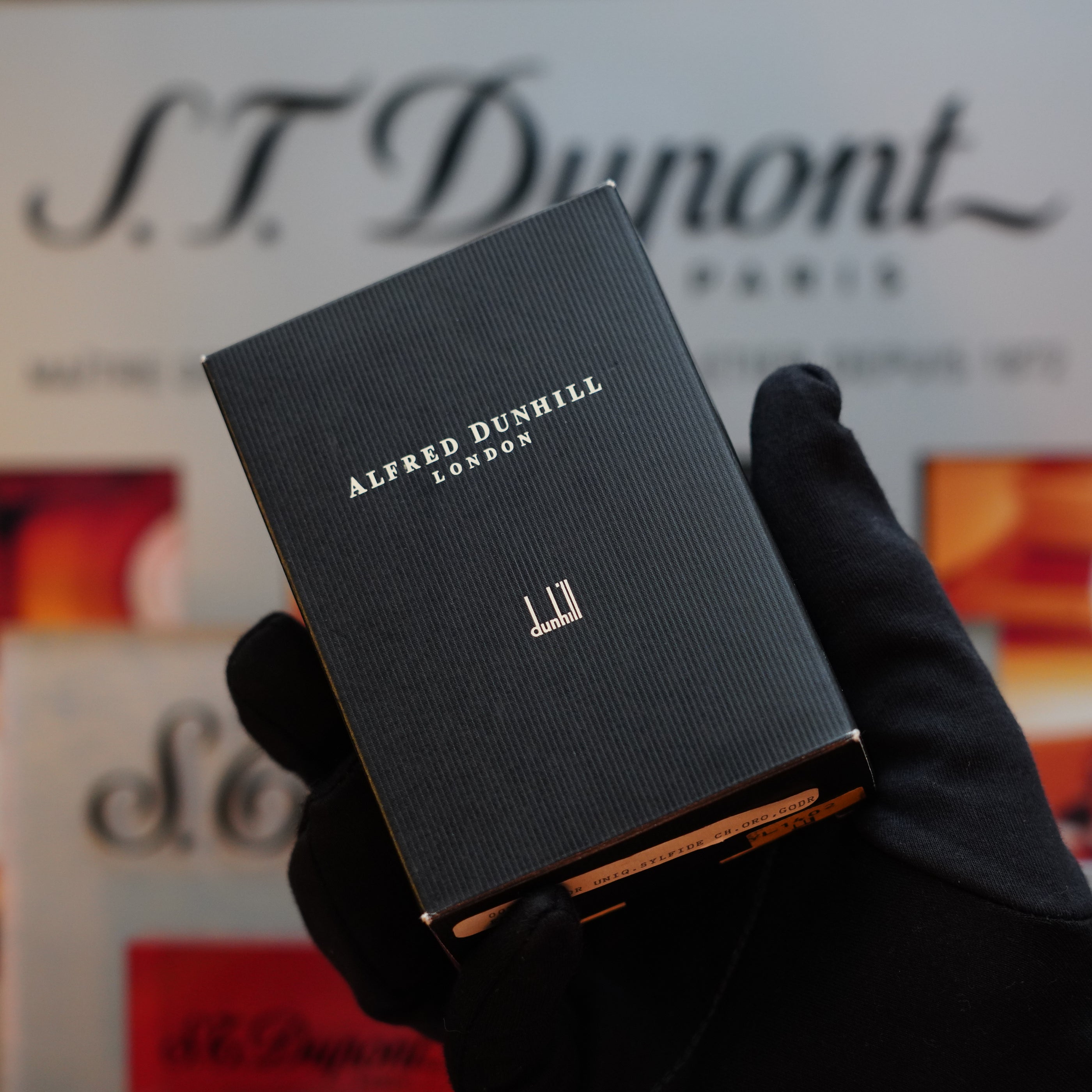 A hand in a black glove holding a small box labeled "Dunhill." The background displays a blurred "S.T. Dupont Paris" logo, evoking the charm of the 1980 Dunhill Unique Sylfide 24k Heavy Gold Natural Lacquer Lighter collection.