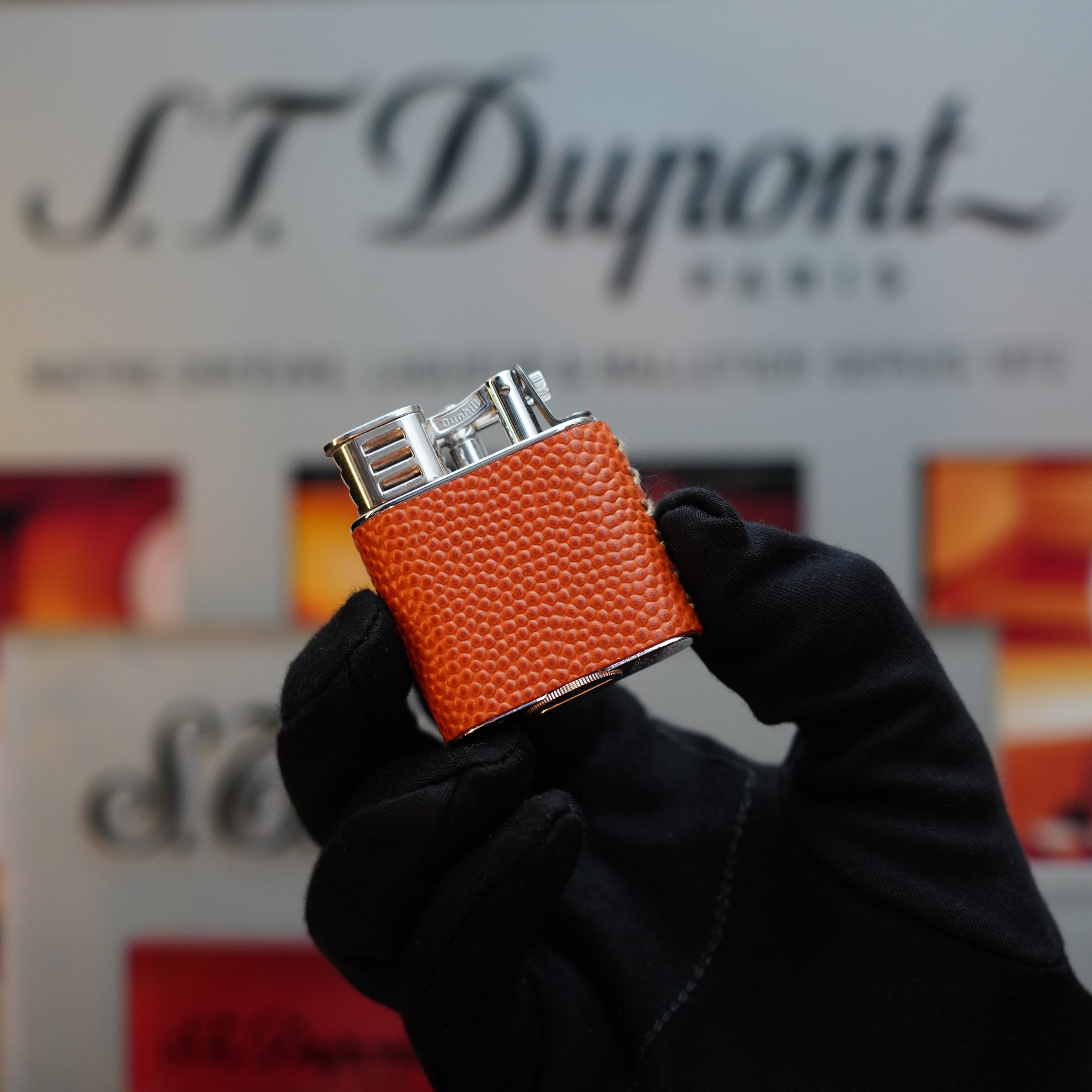 A hand in a black glove holds a Dunhill 1980 Unique Sport Godron lighter with an orange lizard leather body, reminiscent of the Vintage model, against a blurred background featuring S.T. Dupont branding.