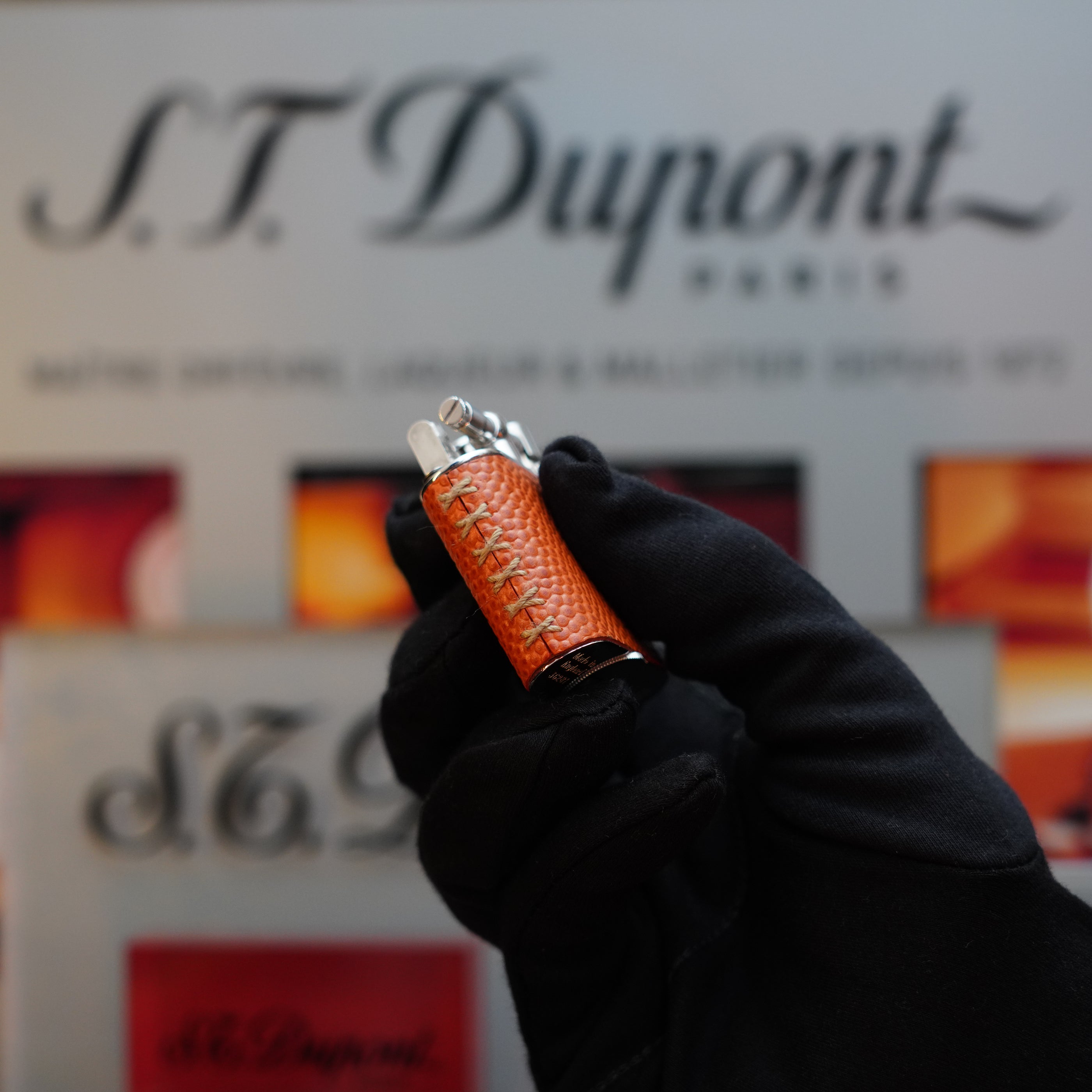 A gloved hand holds an orange textured Dunhill 1980 Unique Sport Godron lighter with a lizard leather body against a blurred background, with the Dunhill logo and text, evoking the prestige of a collector's item from the 80s.