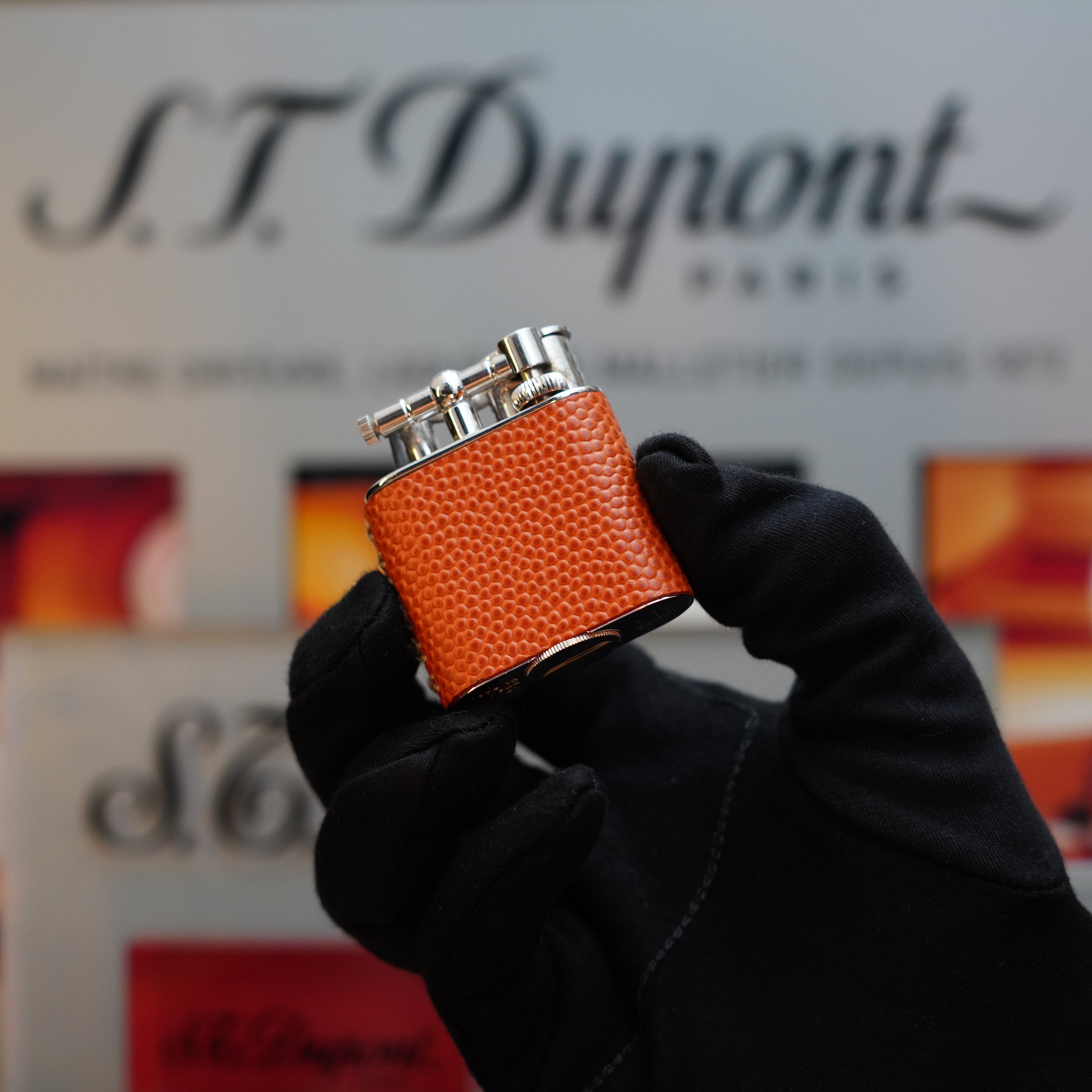 A gloved hand holds an orange leather-wrapped Dunhill Unique Sport Godron lighter against a blurred background displaying the brand's logo—a true collector's item from the 80s.