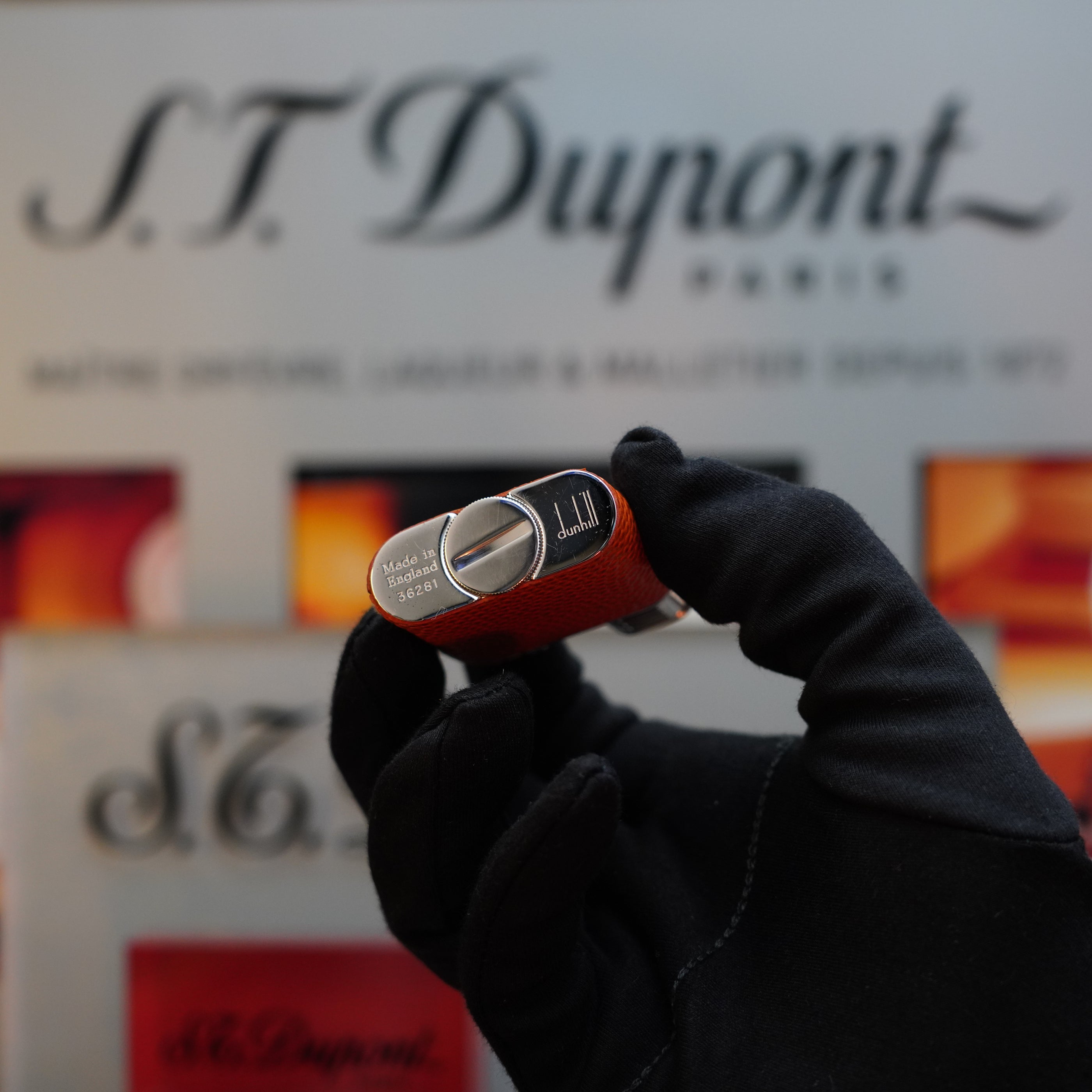 A gloved hand holding a 1980 Dunhill Unique Sport Godron lighter with a lizard orange leather body and benzine fluid functionality, set against a blurred Dunhill branded background—an exquisite collector's item from the 80s.