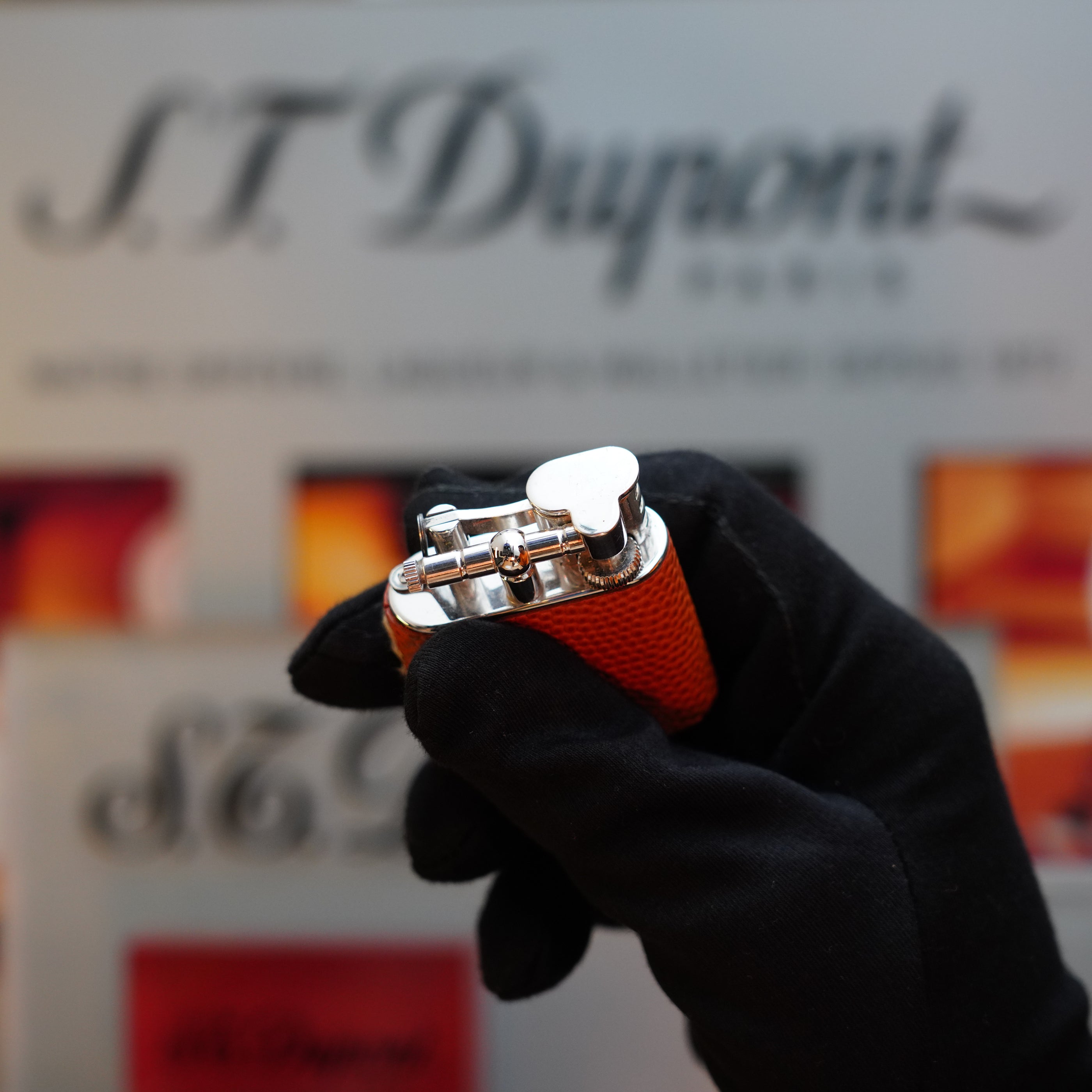 A person wearing a black glove holds an orange Dunhill Unique Sport Godron lighter, featuring a lizard leather body and fueled by benzine, a collector's item from the 1980s, with a background of blurred branded signs.