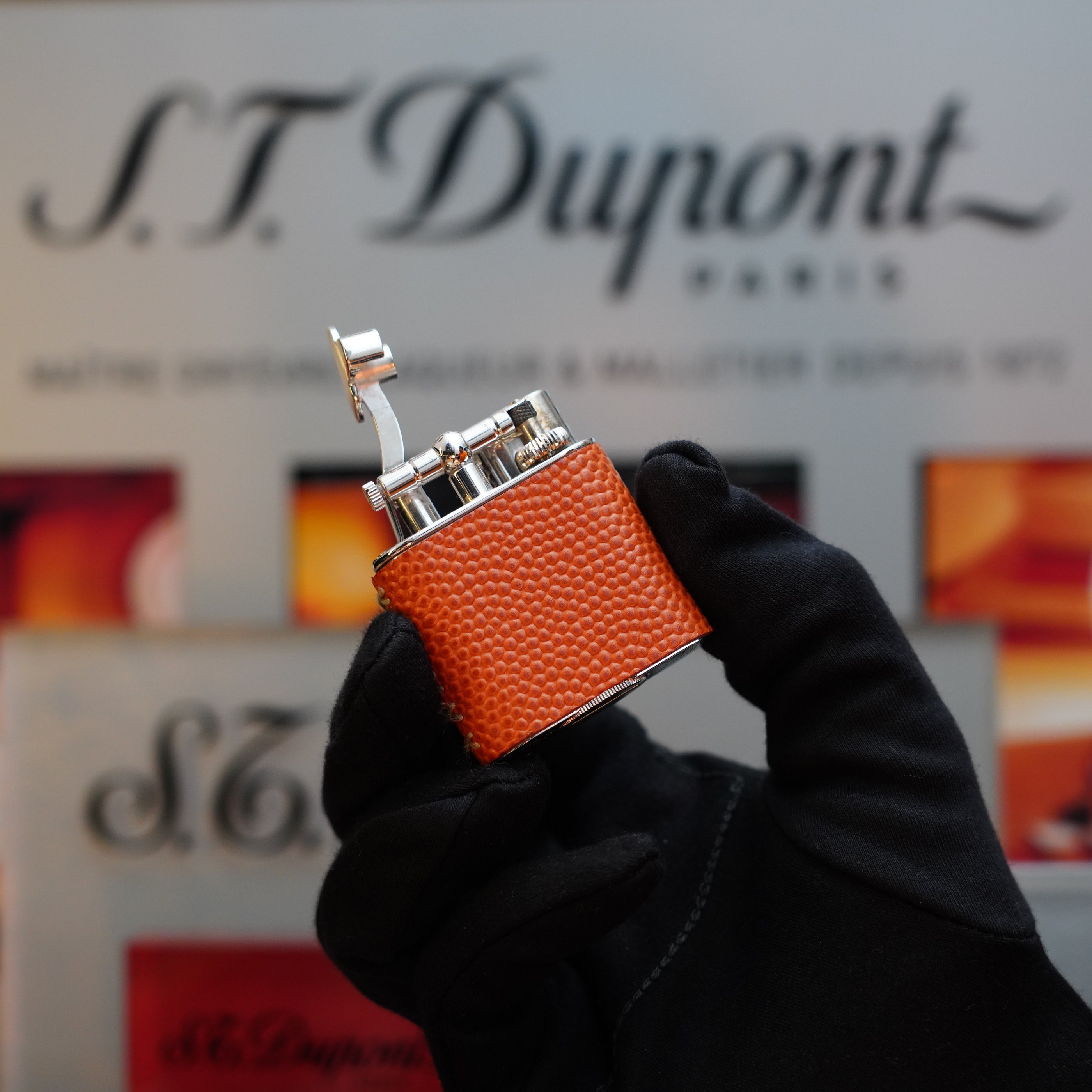 A gloved hand holds an orange textured Dunhill lighter, specifically the 1980 Dunhill Unique Sport Godron With Lizard Orange Leather Body benzine Fluid Lighter, in front of a display featuring multiple S.T. Dupont logos and text, evoking the nostalgia of a collector's item from the 80s.