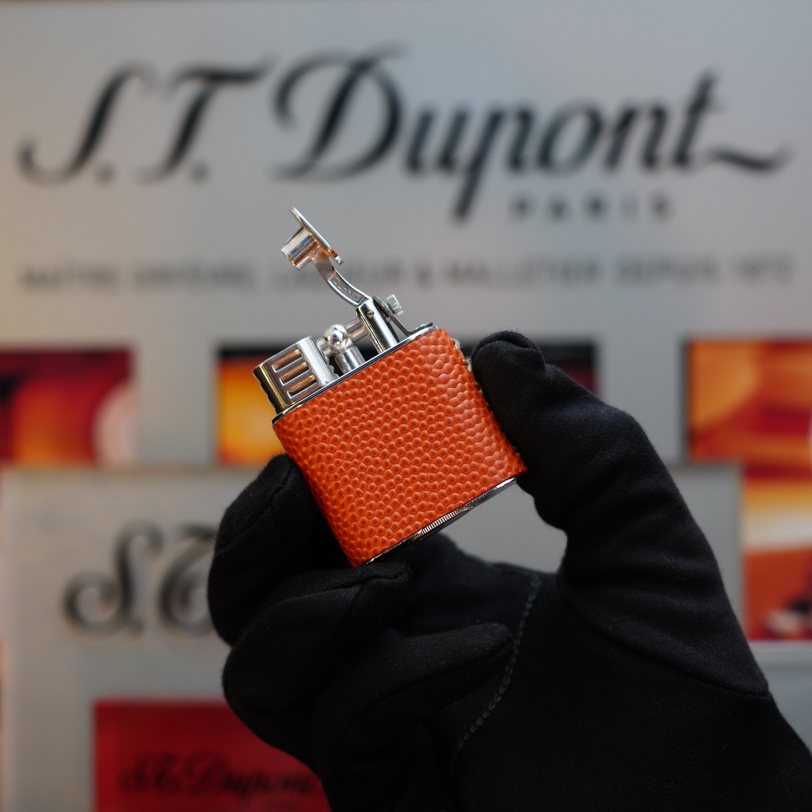 A gloved hand holds a 1980 Dunhill Unique Sport Godron lighter with an orange lizard leather body in front of a display featuring the Dunhill brand name, evoking memories of vintage elegance.