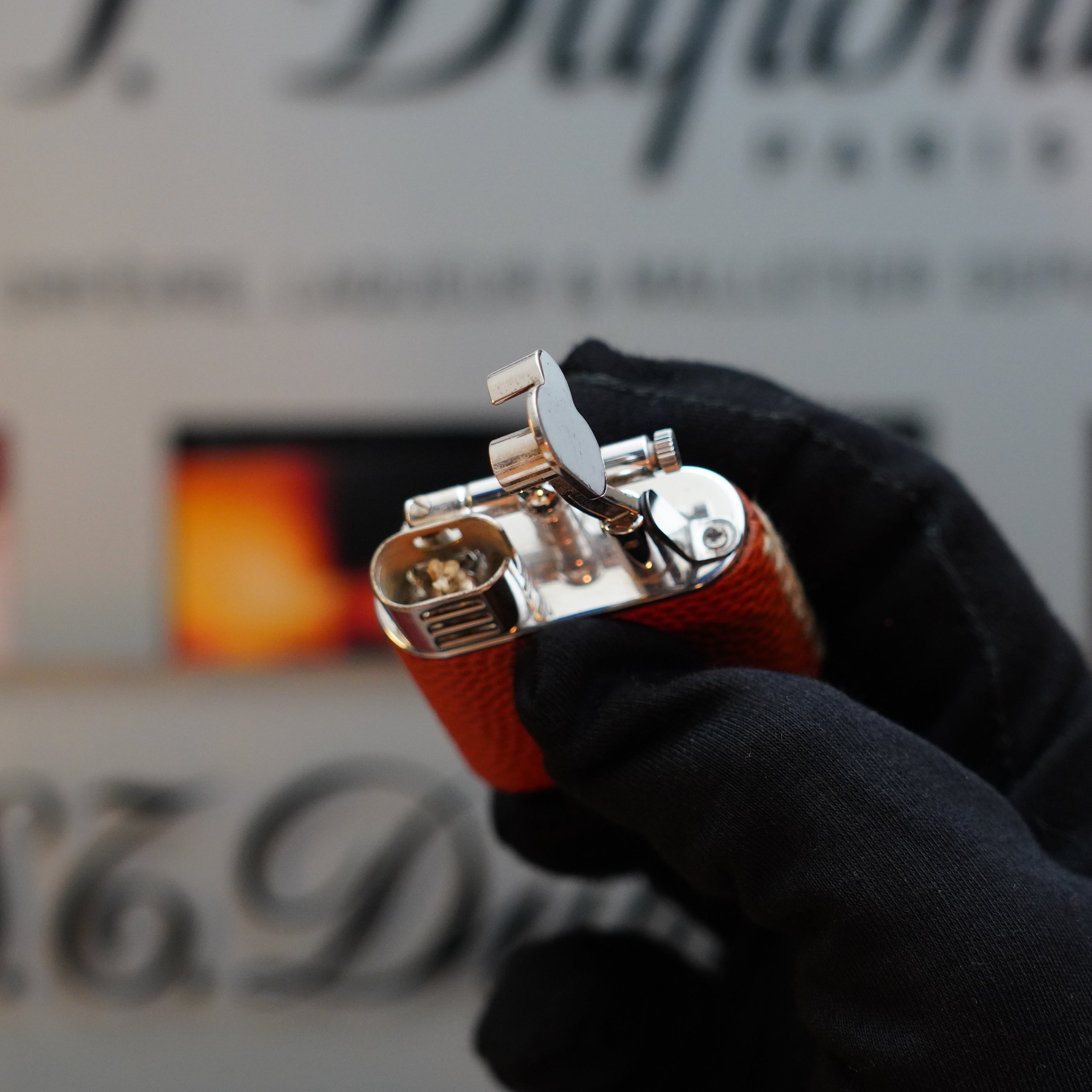 A gloved hand holds a silver and orange lizard leather lighter, the 1980 Dunhill Unique Sport Godron, cherished as a collector's item. The background is slightly blurred with some text and colors visible.