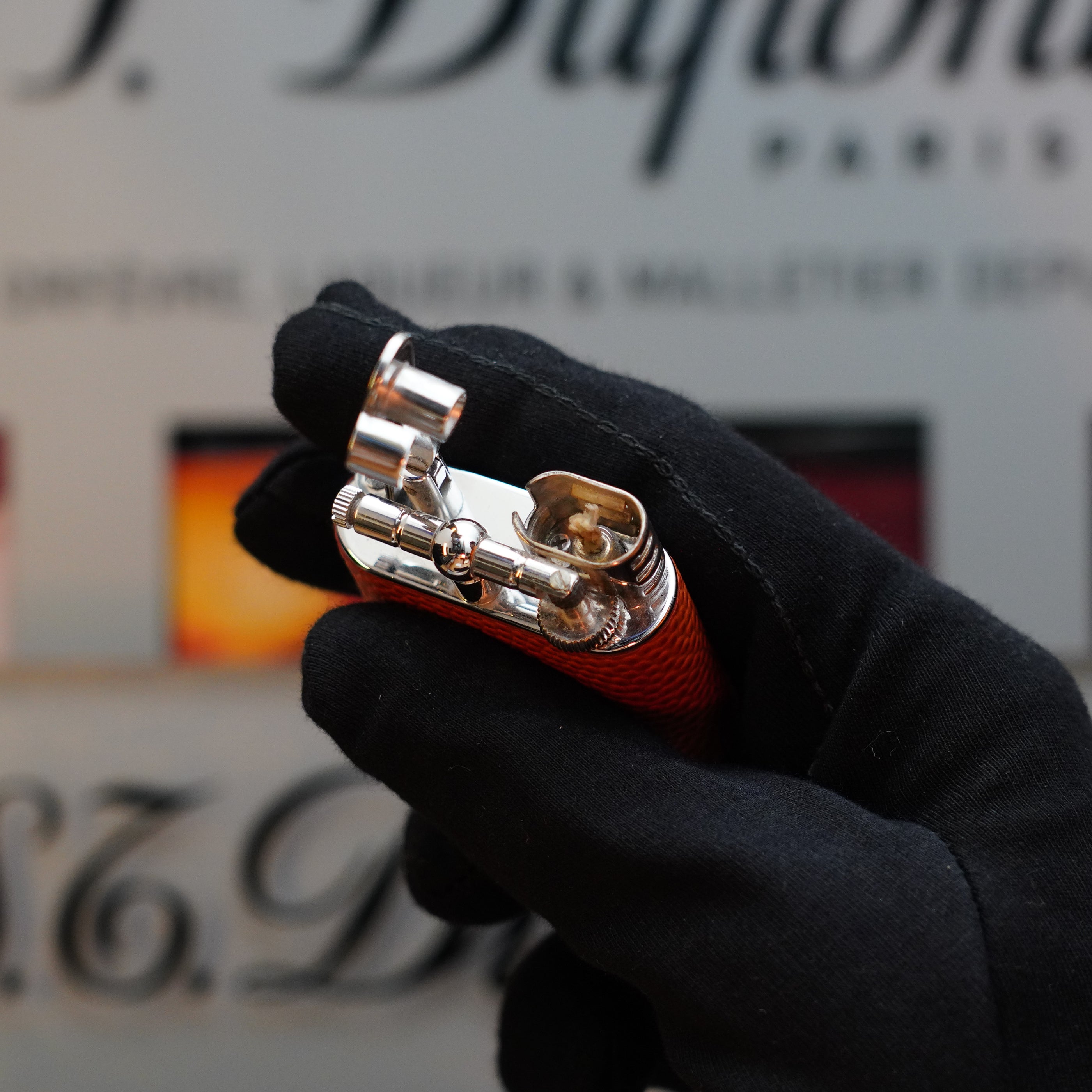 A gloved hand holds an orange and silver 1980 Dunhill Unique Sport Godron with Lizard Orange Leather Body benzine fluid lighter, with the cap open, ready for use. The background is blurred, showcasing partial, out-of-focus text. This elegant piece from the 80s is not just functional but also a collector's item.