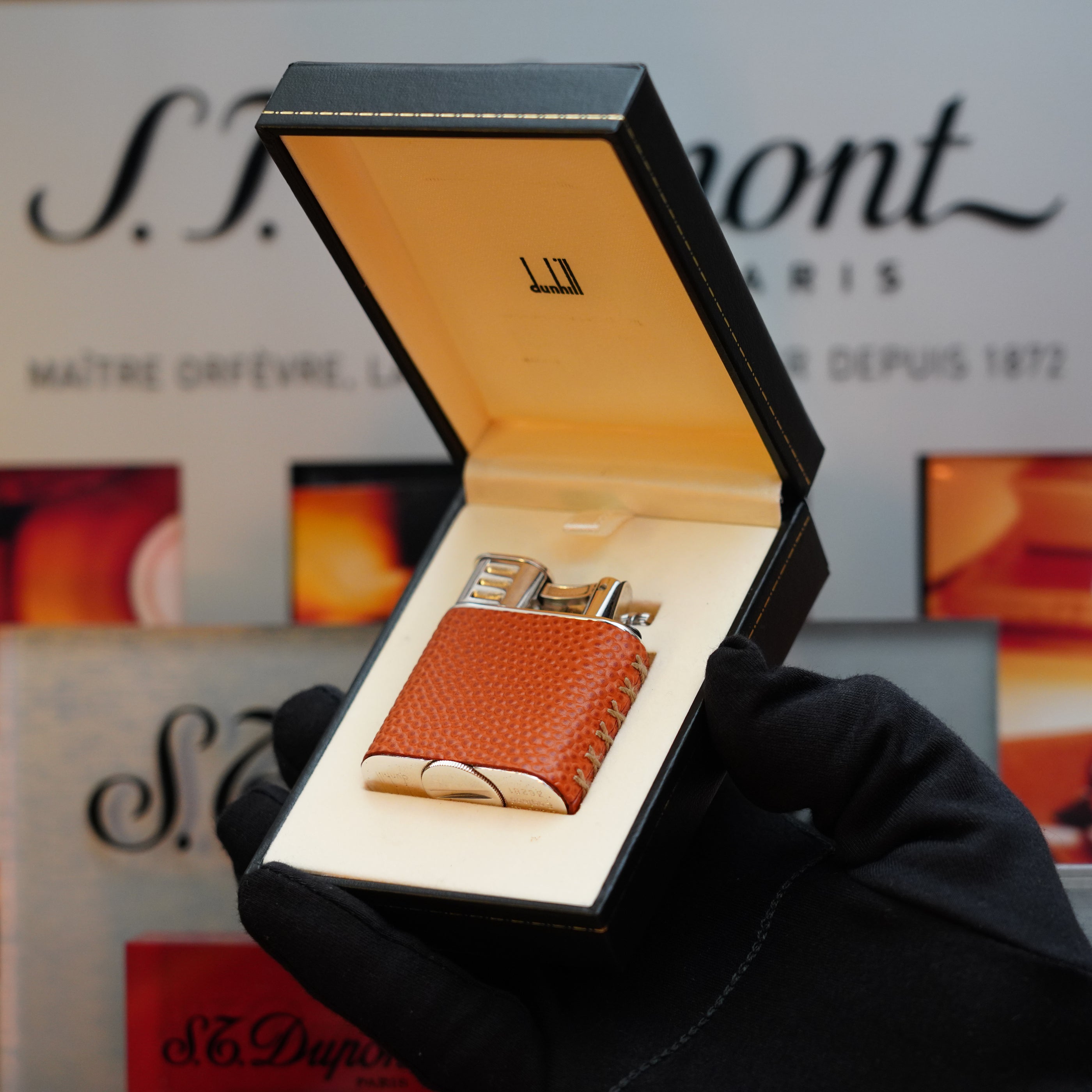 A gloved hand holds an open black box displaying a 1980 Dunhill Unique Sport Godron lighter with an orange textured lizard leather body. The background features blurred text and images related to S.T. Dupont, showcasing a collector's item from the 80s.