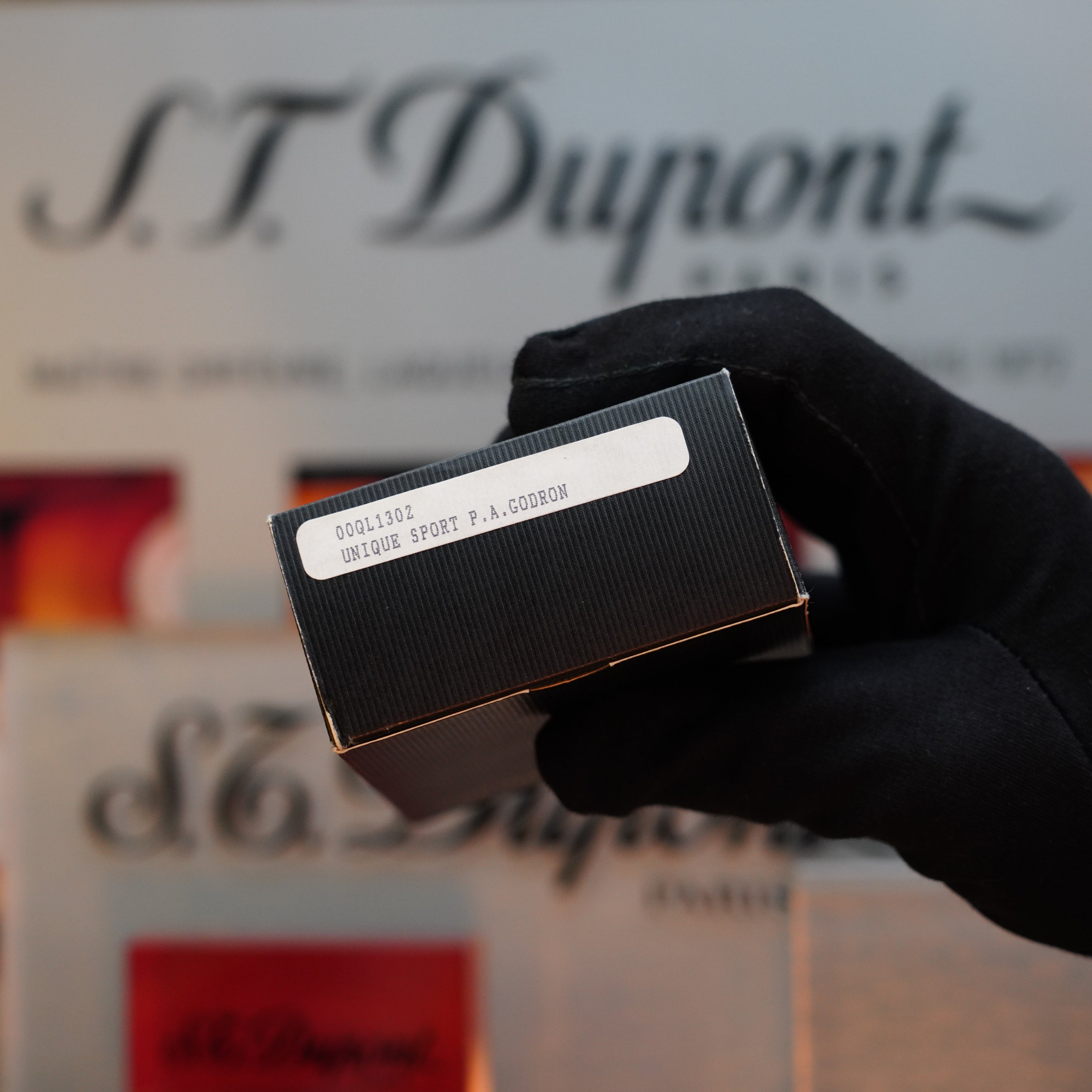 A gloved hand holds a black box labeled "1980 Dunhill Unique Sport Godron," revealing a collector's item from the 80s with Dunhill branding visible in the background.