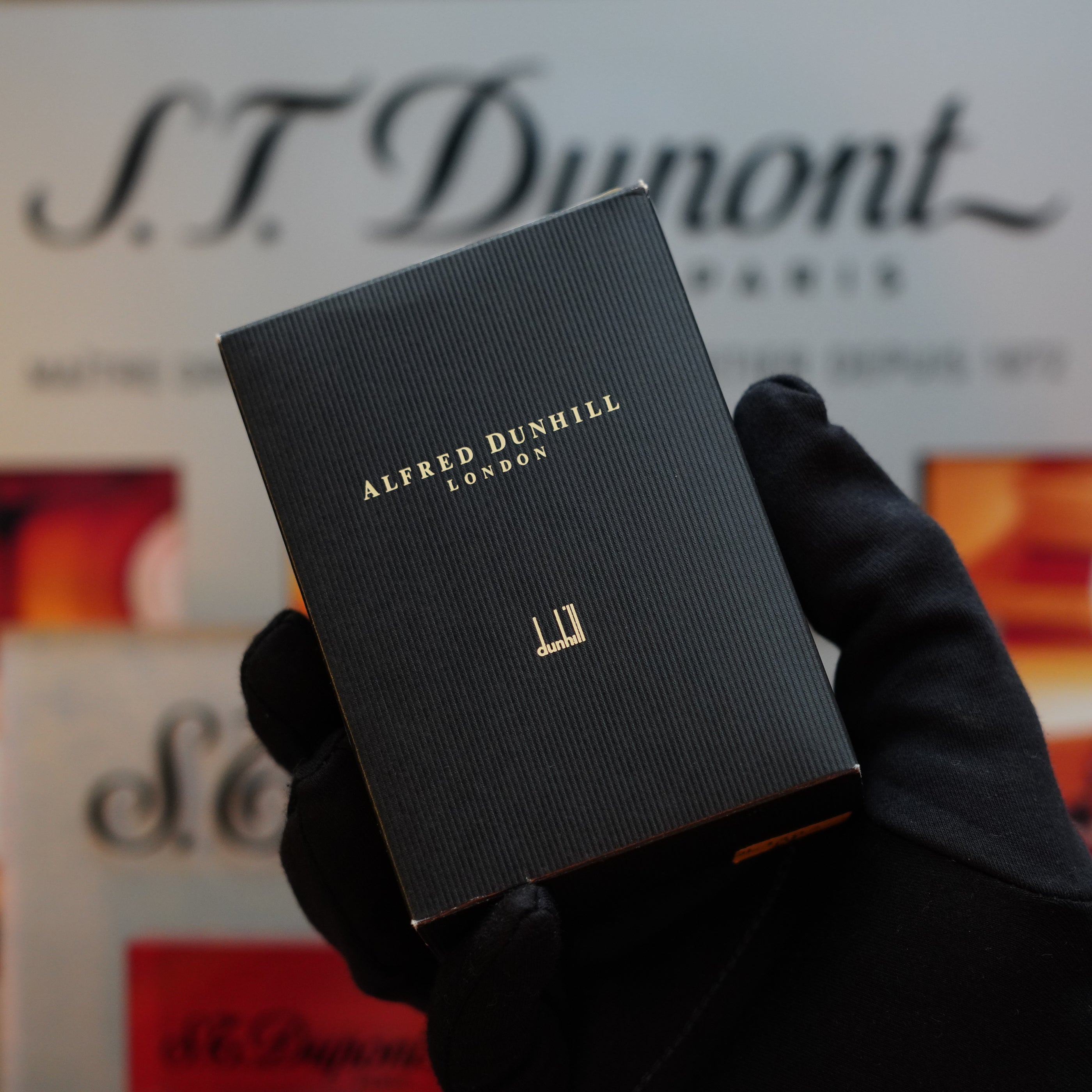 A gloved hand holds a small black box with gold text that reads "Dunhill." This vintage 1980 Dunhill Unique Sport Godron, featuring a Lizard Orange Leather Body and using benzine fluid, is set against the blurred logo and text of S.T. Dupont Paris—a true collector's item from the 80s.