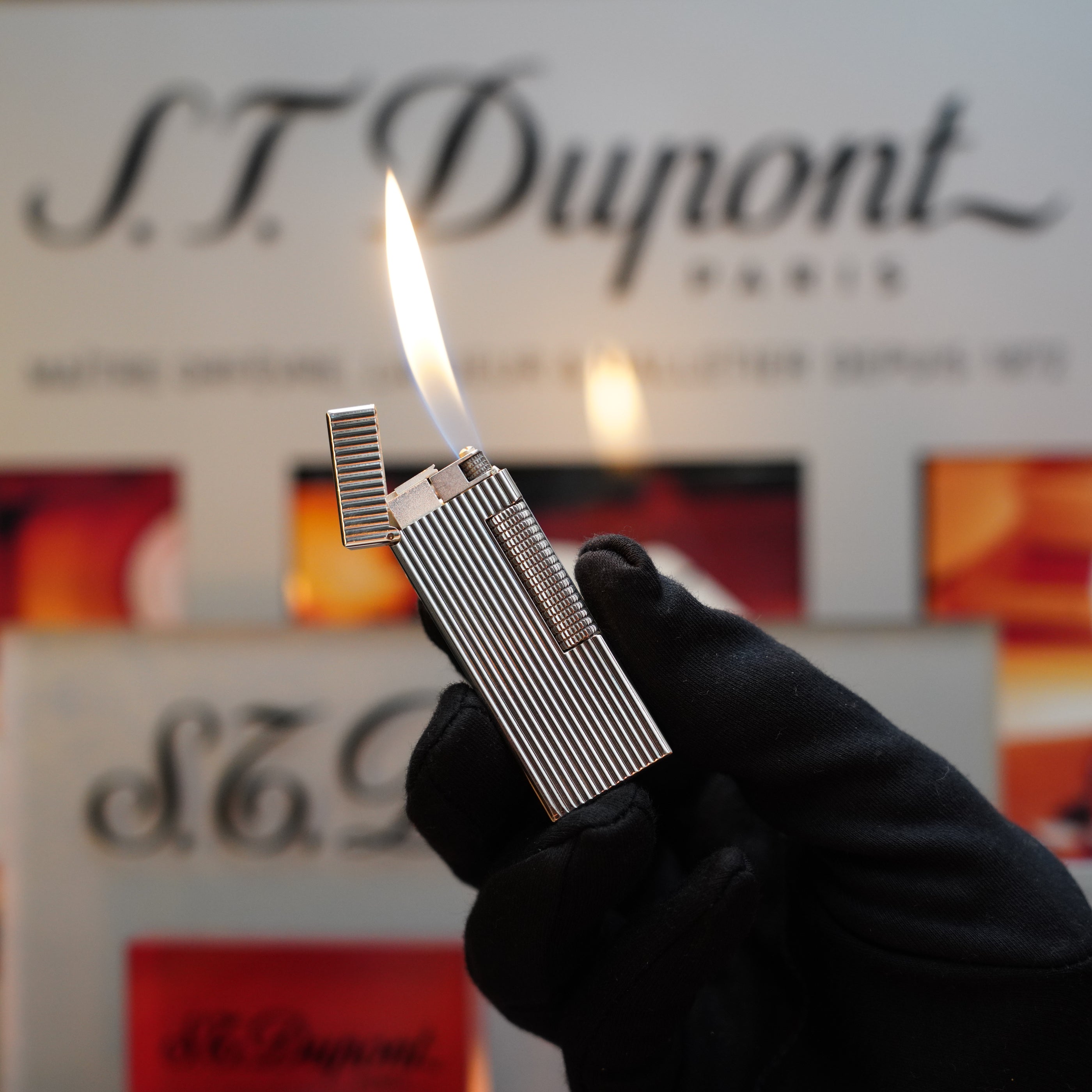 A gloved hand holds a lit 1970 Dunhill Rollagas Vertical Line Heavy Silver Finish Lighter in front of an S.T. Dupont Paris background display, capturing the essence of a true collector's item.