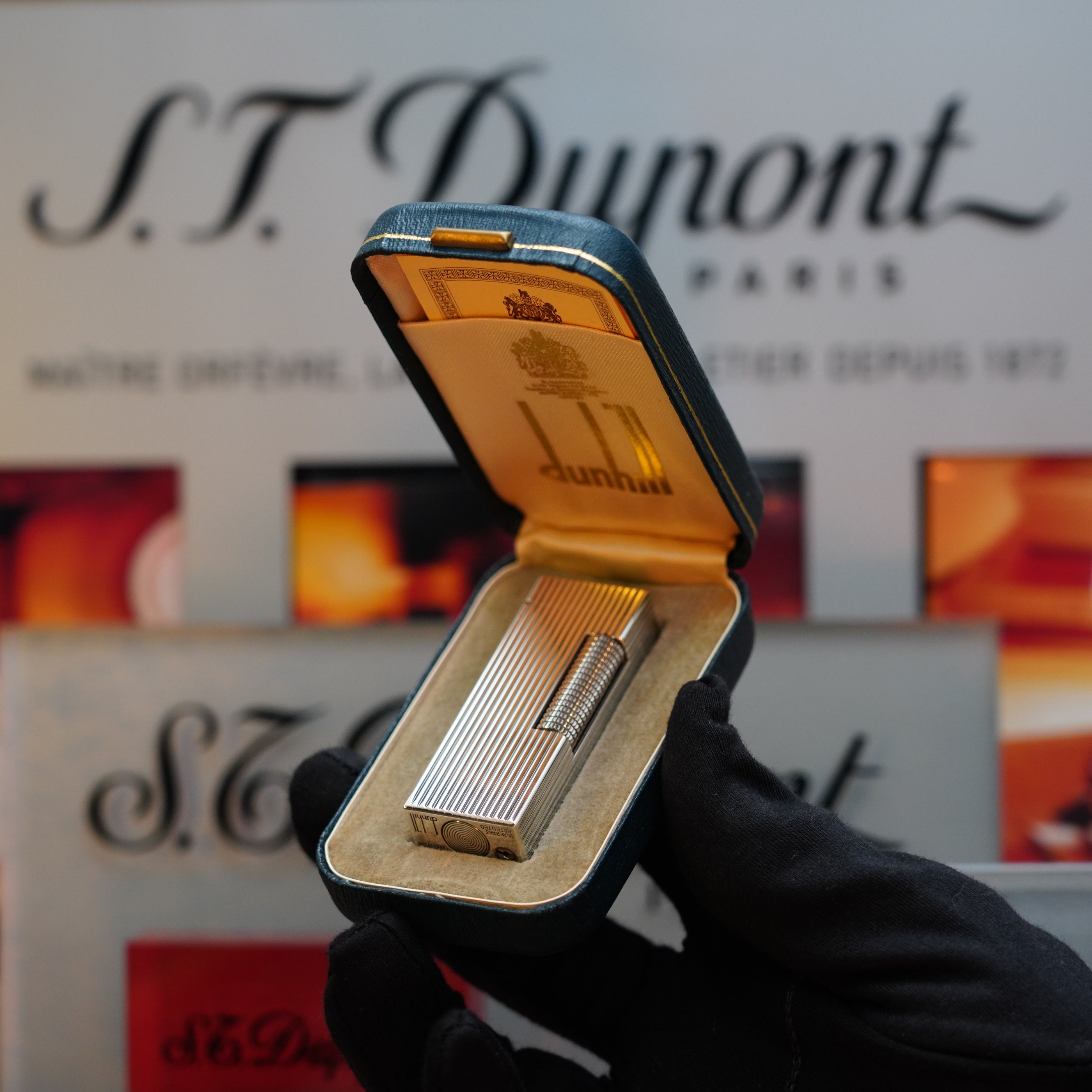 A gloved hand holds an open box displaying a silver 1970 Dunhill Rollagas Vertical Line Heavy Silver Finish Lighter, against a blurred background featuring the Dunhill logo—a true collector's item.