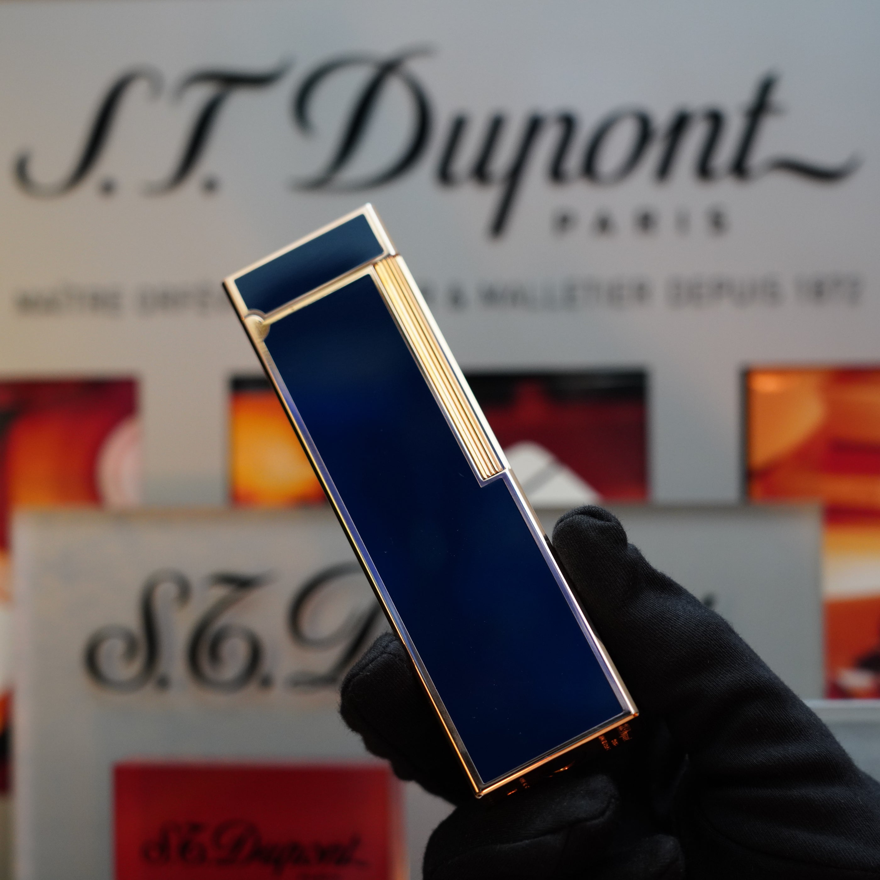 A person wearing a black glove holds a 1970 St Dupont Natural Rare Blue Lacquer Table Lighter with a heavy gold finish. The background features S.T. Dupont branding.