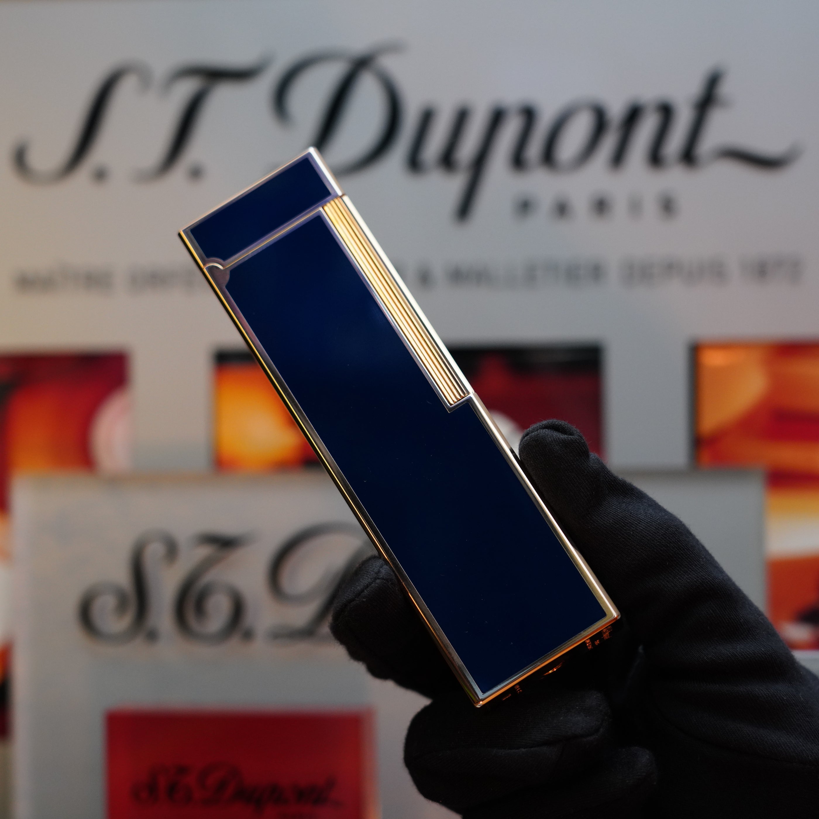 A gloved hand holds a blue and gold 1970 St Dupont Natural rare Blue Lacquer Table Lighter Heavy Gold Finish, with the S.T. Dupont logo visible in the background.