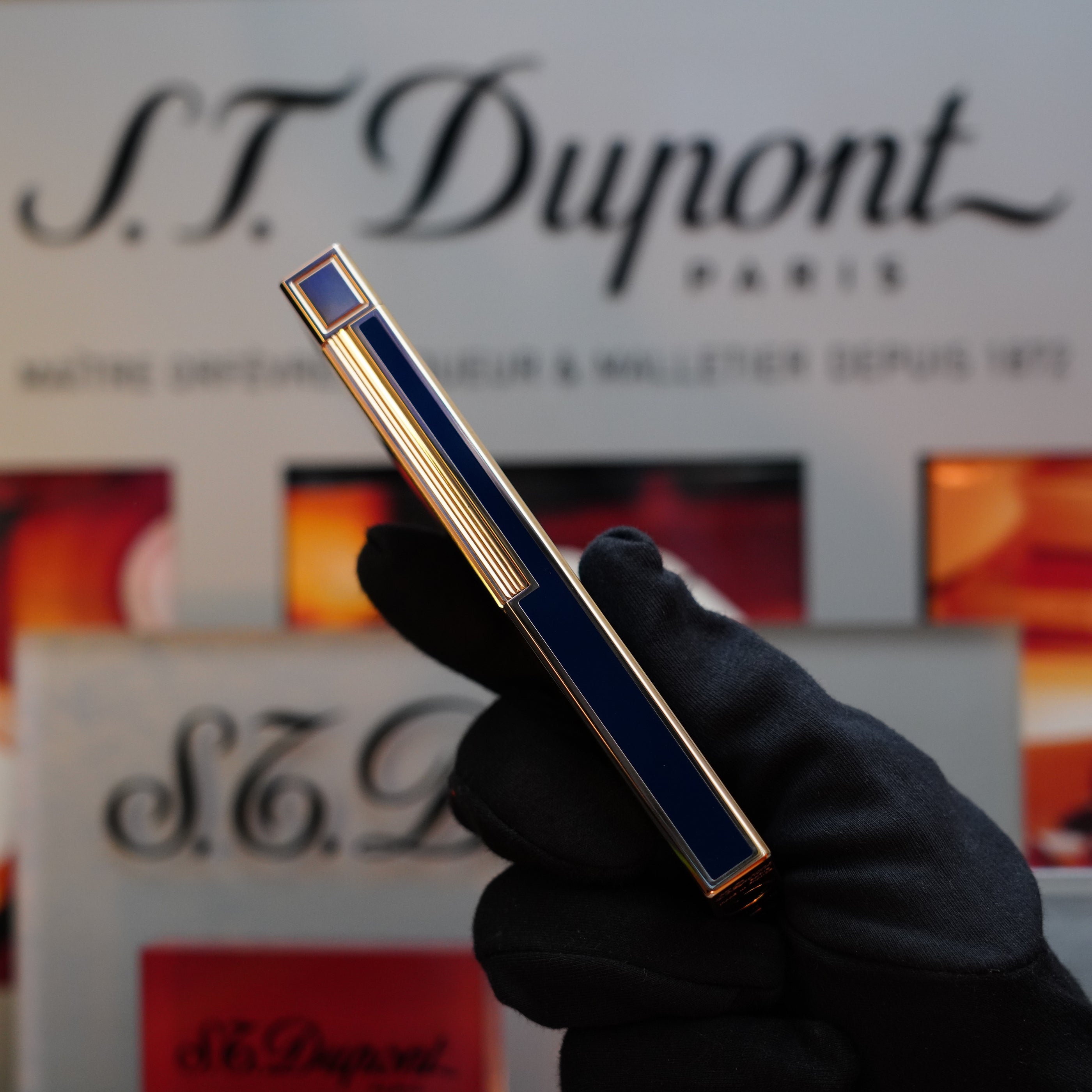 A gloved hand holds a 1970 S.T. Dupont Natural rare Blue Lacquer Table Lighter with a heavy gold finish, reminiscent of the sophisticated style seen in the Ligne 1 Series, against a blurred background featuring S.T. Dupont branding.