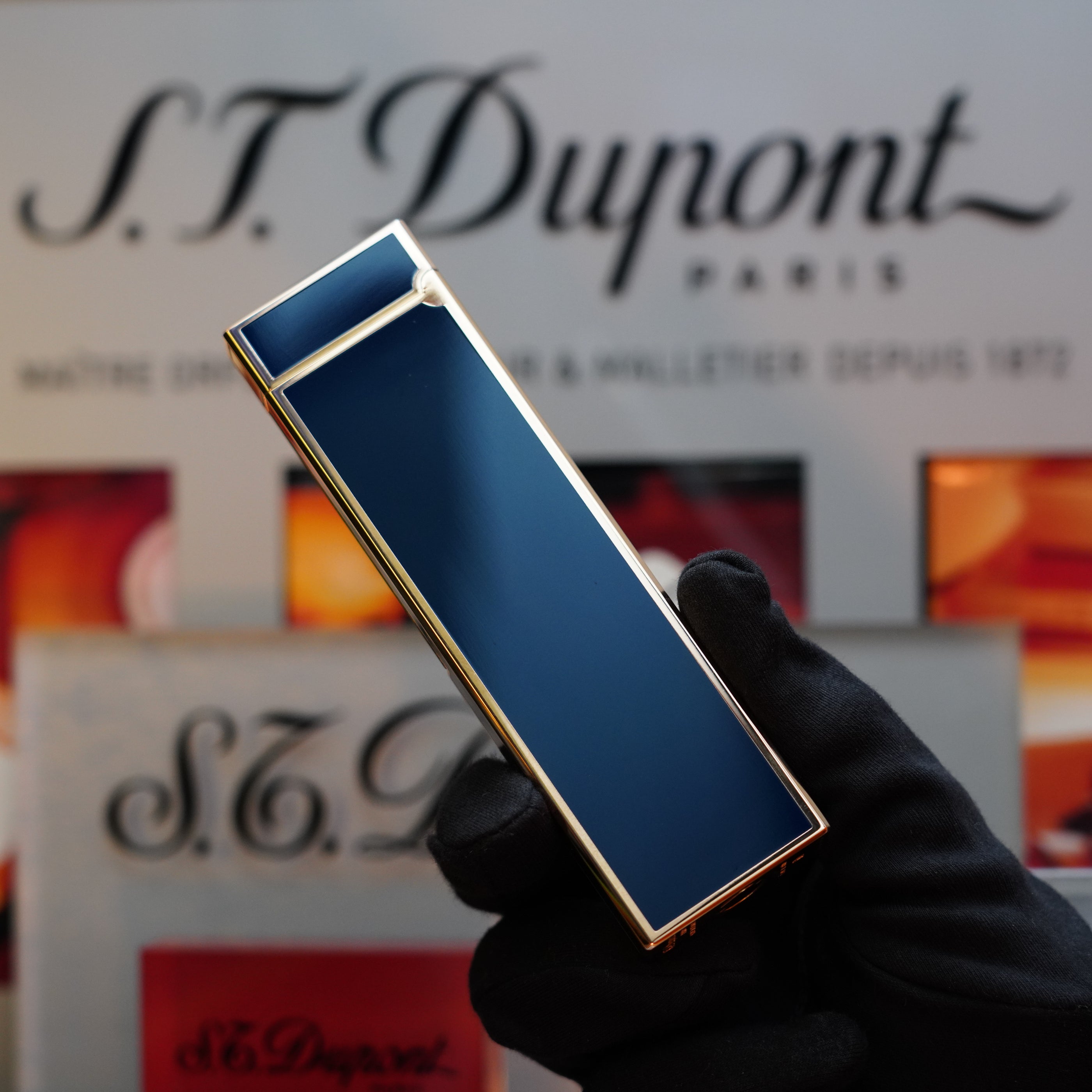 A gloved hand holds a rectangular 1970 St Dupont Natural rare Blue Lacquer Table Lighter with a Heavy Gold Finish, with a blurred S.T. Dupont Paris logo in the background.