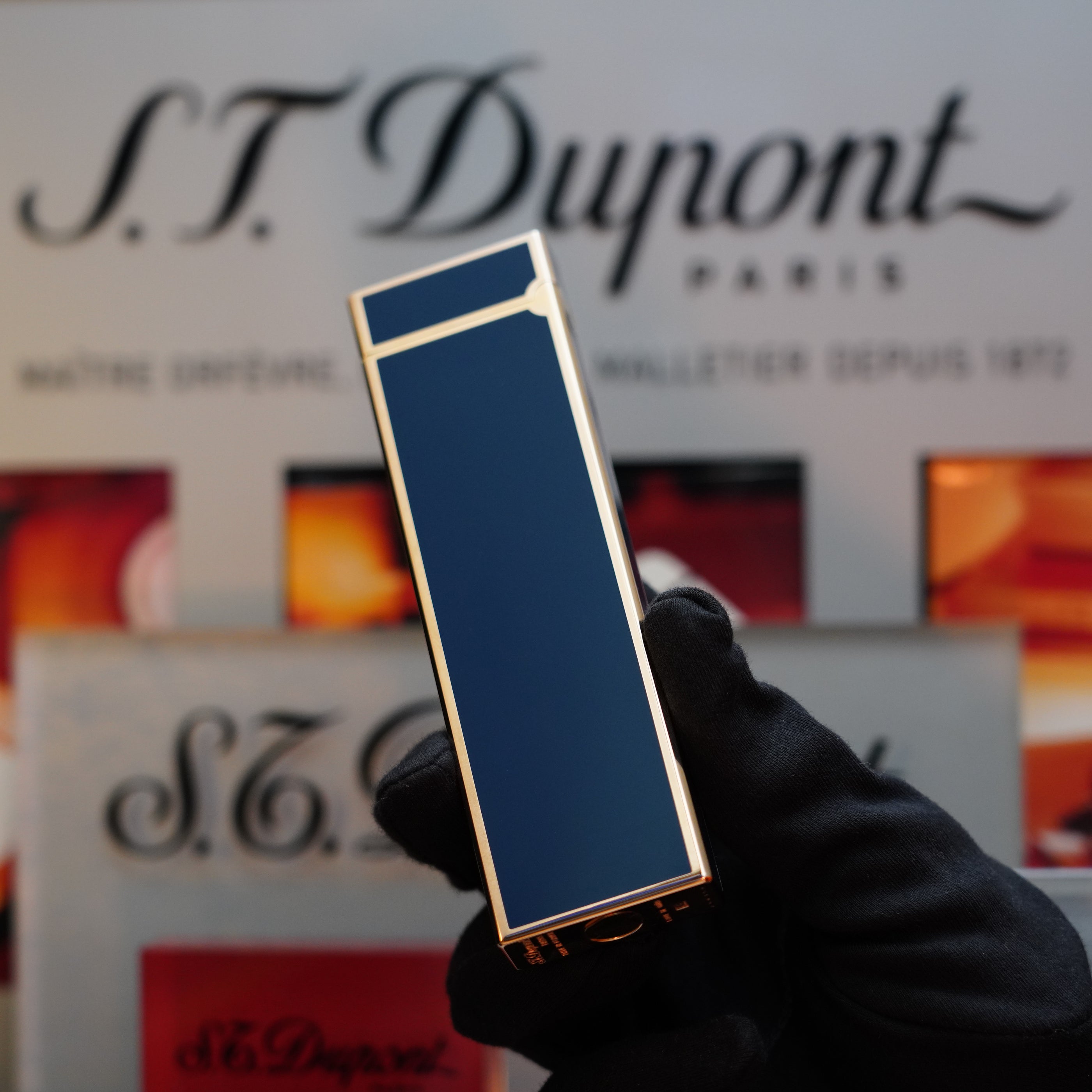 A person wearing a black glove holds a 1970 S.T. Dupont Natural rare Blue Lacquer Table Lighter with a Heavy Gold Finish, showcasing its elegance against a background featuring S.T. Dupont branding and text.