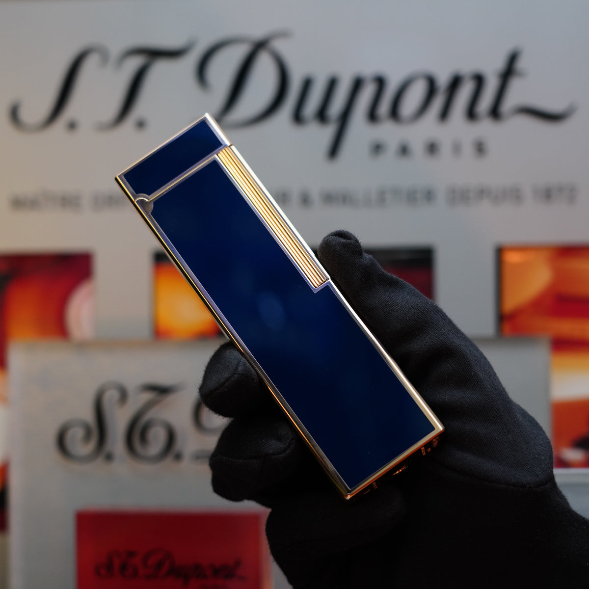 A gloved hand holds a sleek blue rectangular object in front of an S.T. Dupont Paris sign, showcasing a rare 1970 St Dupont Natural Blue Lacquer Table Lighter with a Heavy Gold Finish.
