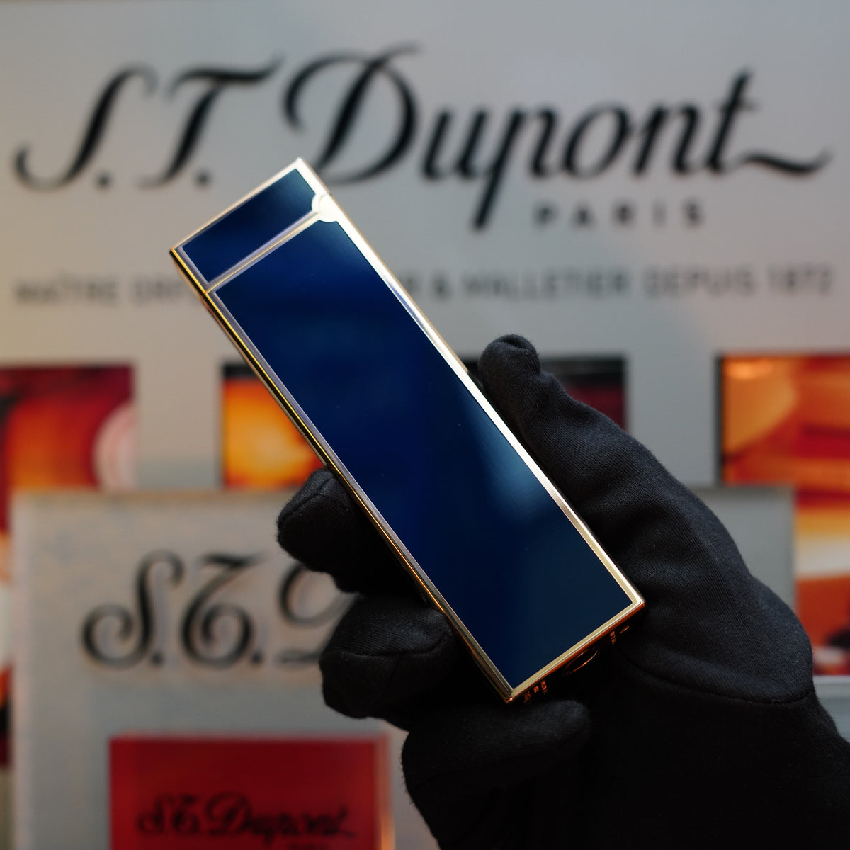 A gloved hand holding a rectangular, dark blue, gold-trimmed 1970 St Dupont Natural rare Blue Lacquer Table Lighter Heavy Gold Finish from S.T. Dupont in front of a blurred S.T. Dupont Paris logo backdrop.