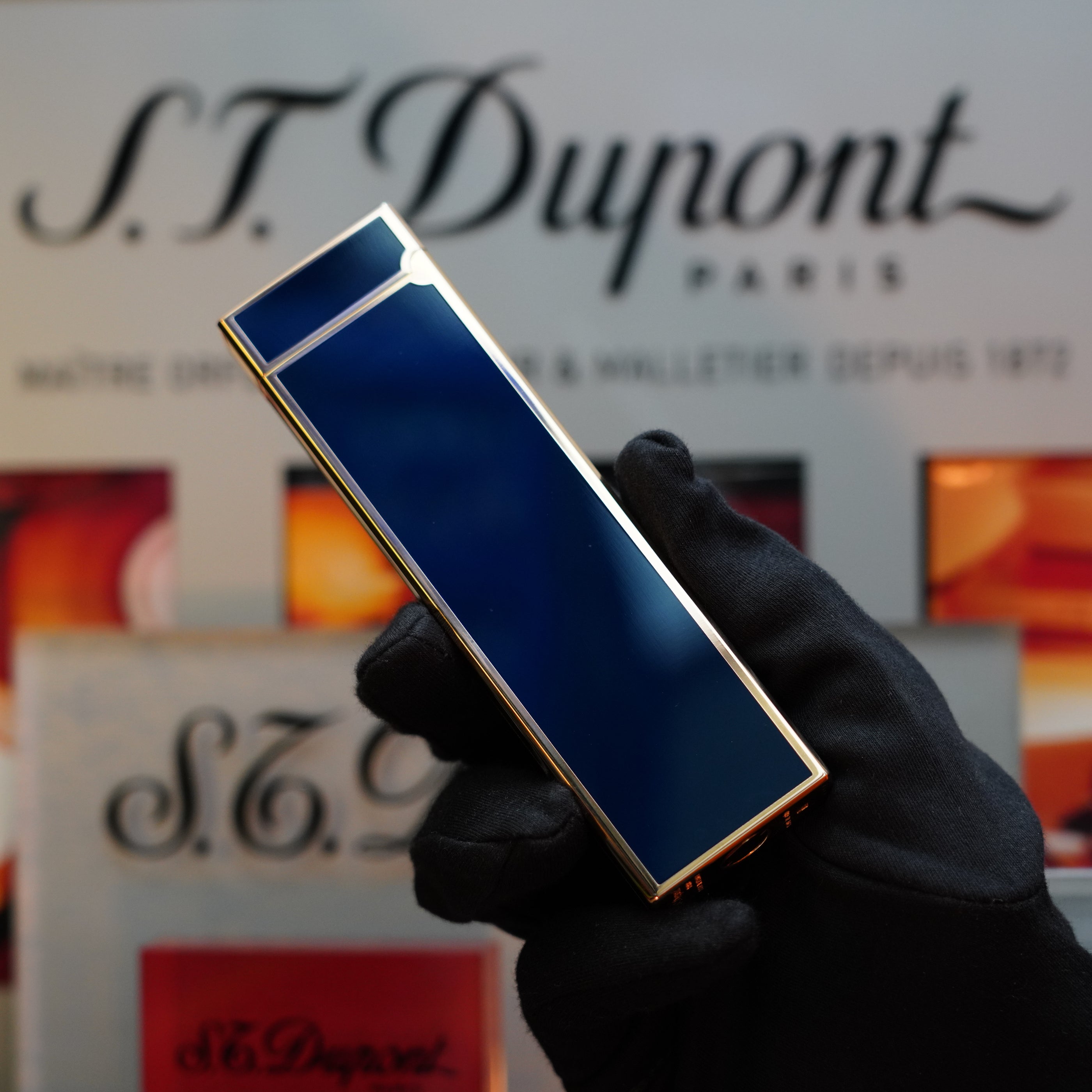 A gloved hand holding a rectangular, dark blue, gold-trimmed 1970 St Dupont Natural rare Blue Lacquer Table Lighter Heavy Gold Finish from S.T. Dupont in front of a blurred S.T. Dupont Paris logo backdrop.