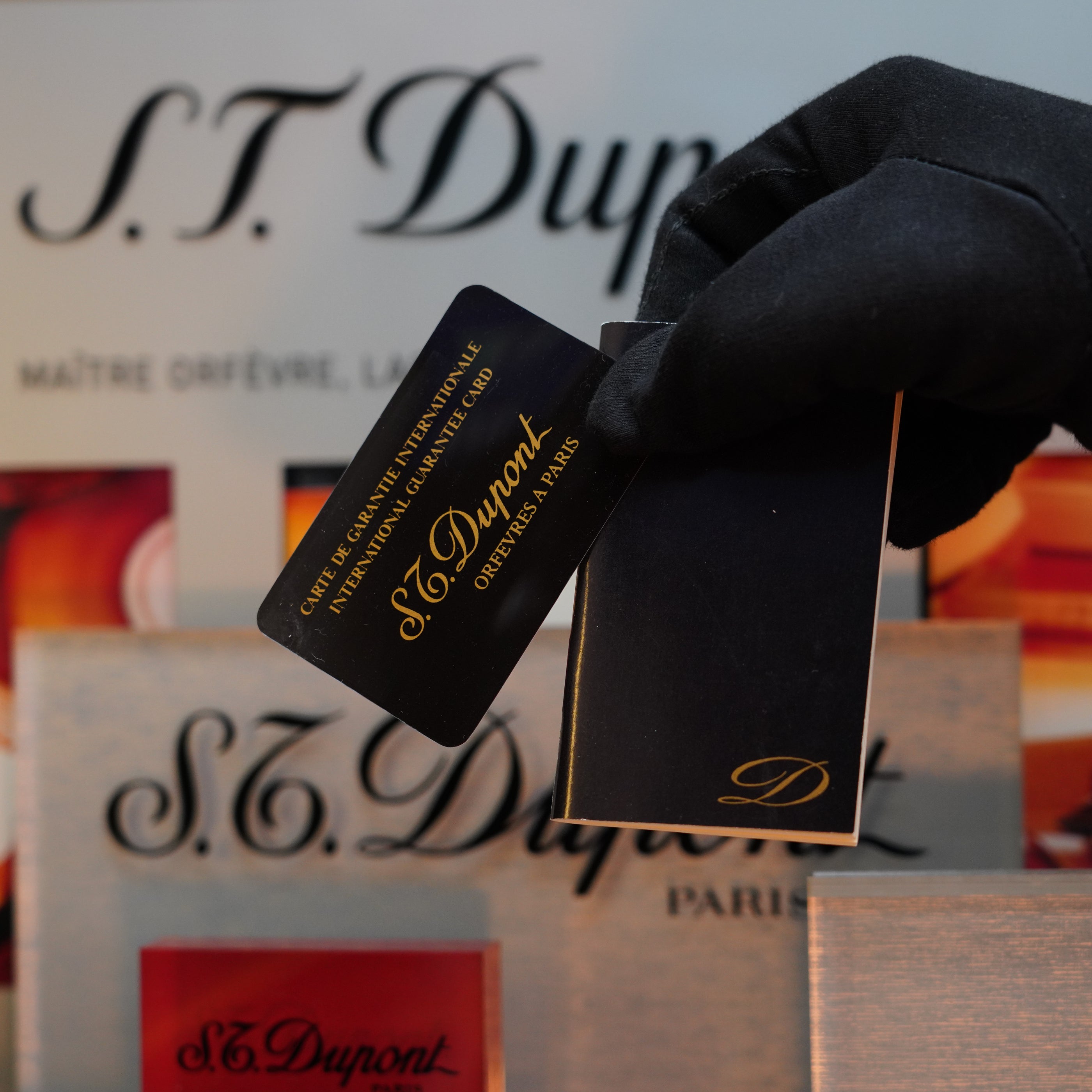A gloved hand holds an S.T. Dupont certificate card and a booklet, while various S.T. Dupont products, including the 1970 St Dupont Natural rare Blue Lacquer Table Lighter Heavy Gold Finish, are displayed in the background.