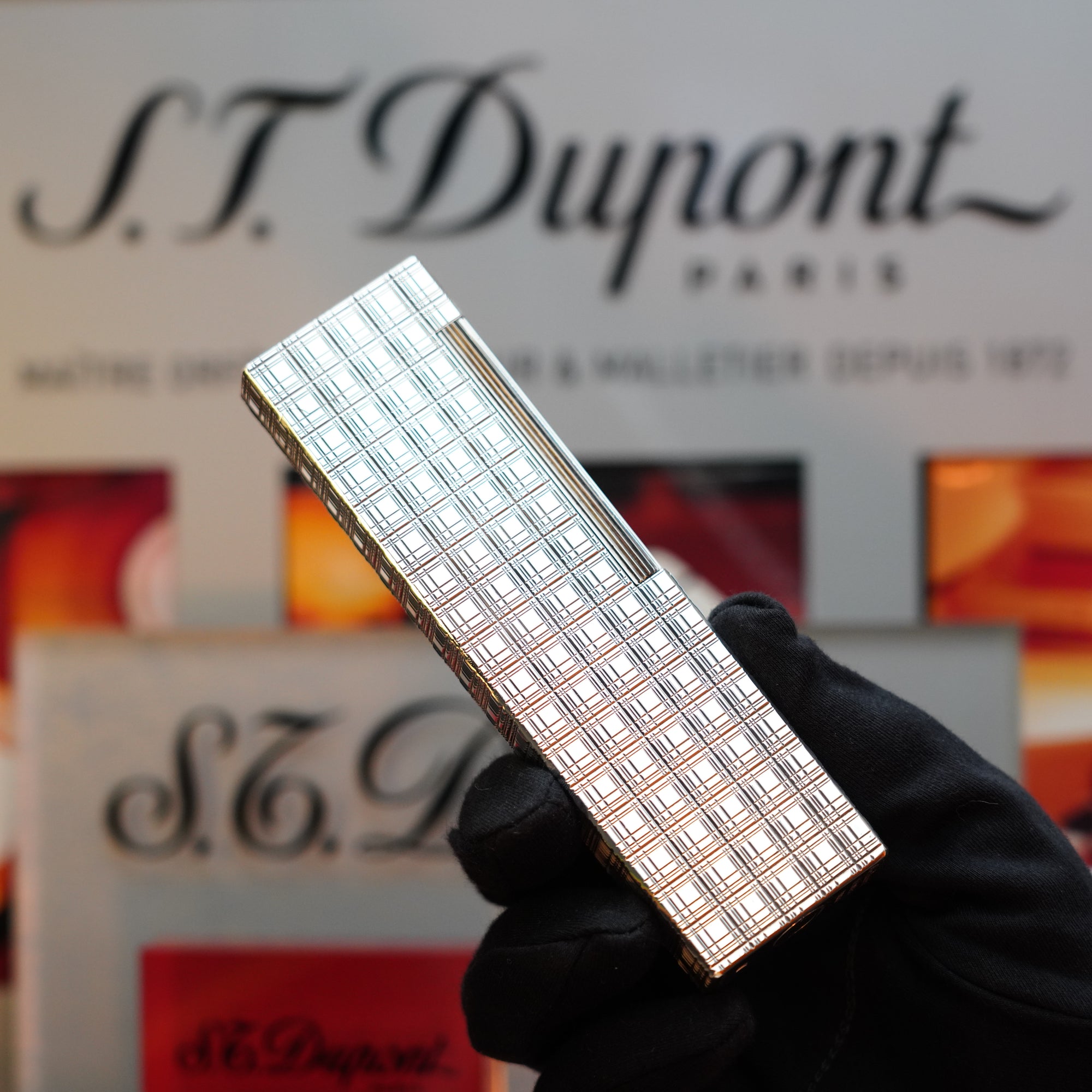 A gloved hand holds a silver patterned S.T. Dupont lighter with blurred S.T. Dupont branding in the background, underscoring that this collector's item is as exquisite as the 1960 HERMES Table Lighter with a St Dupont Platinum Finish in a rare pattern.