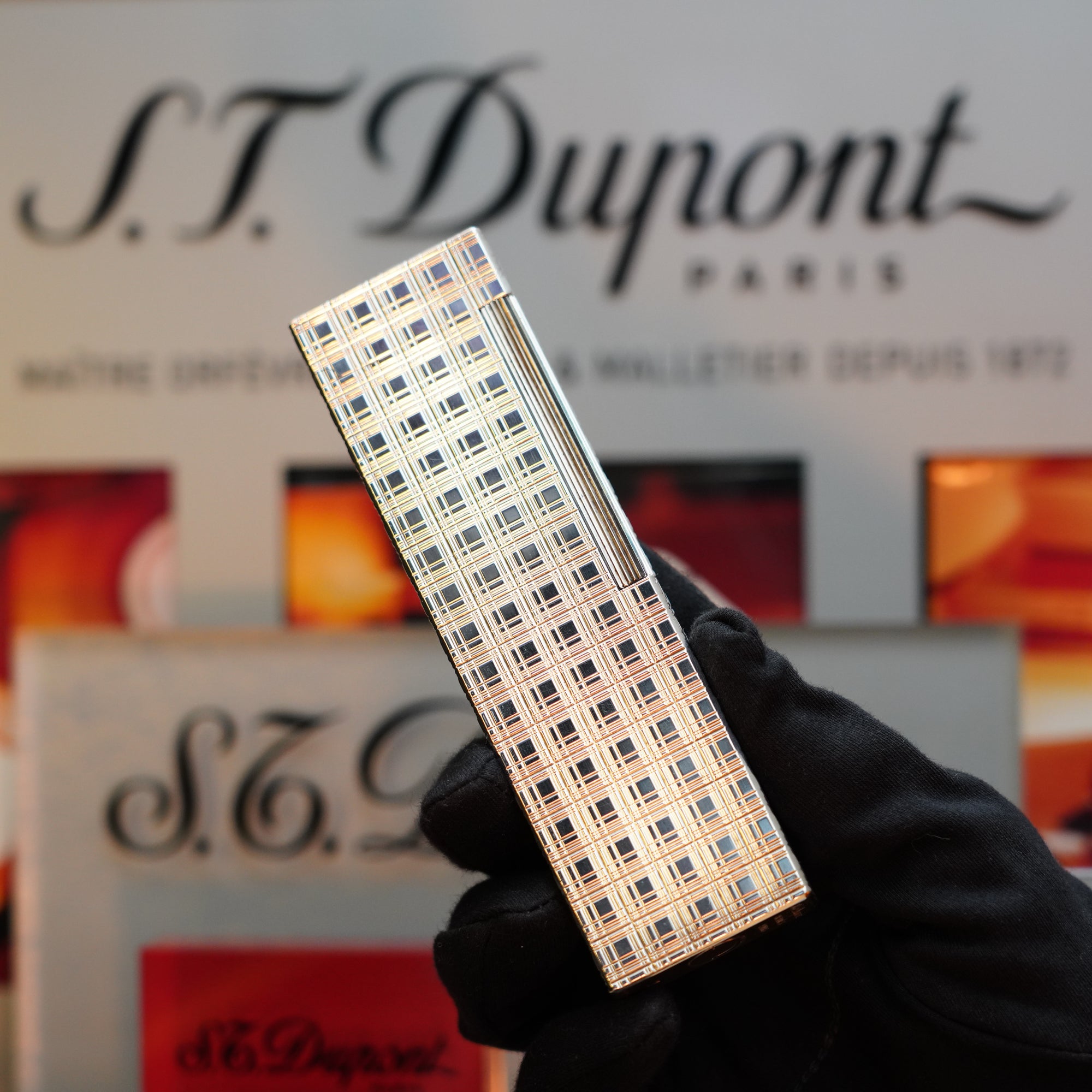 A gloved hand holds the 1960 HERMES Table Lighter with a rare checkered pattern and St Dupont Platinum Finish in front of an S.T. Dupont signage, showcasing a collector's item from the renowned brand.