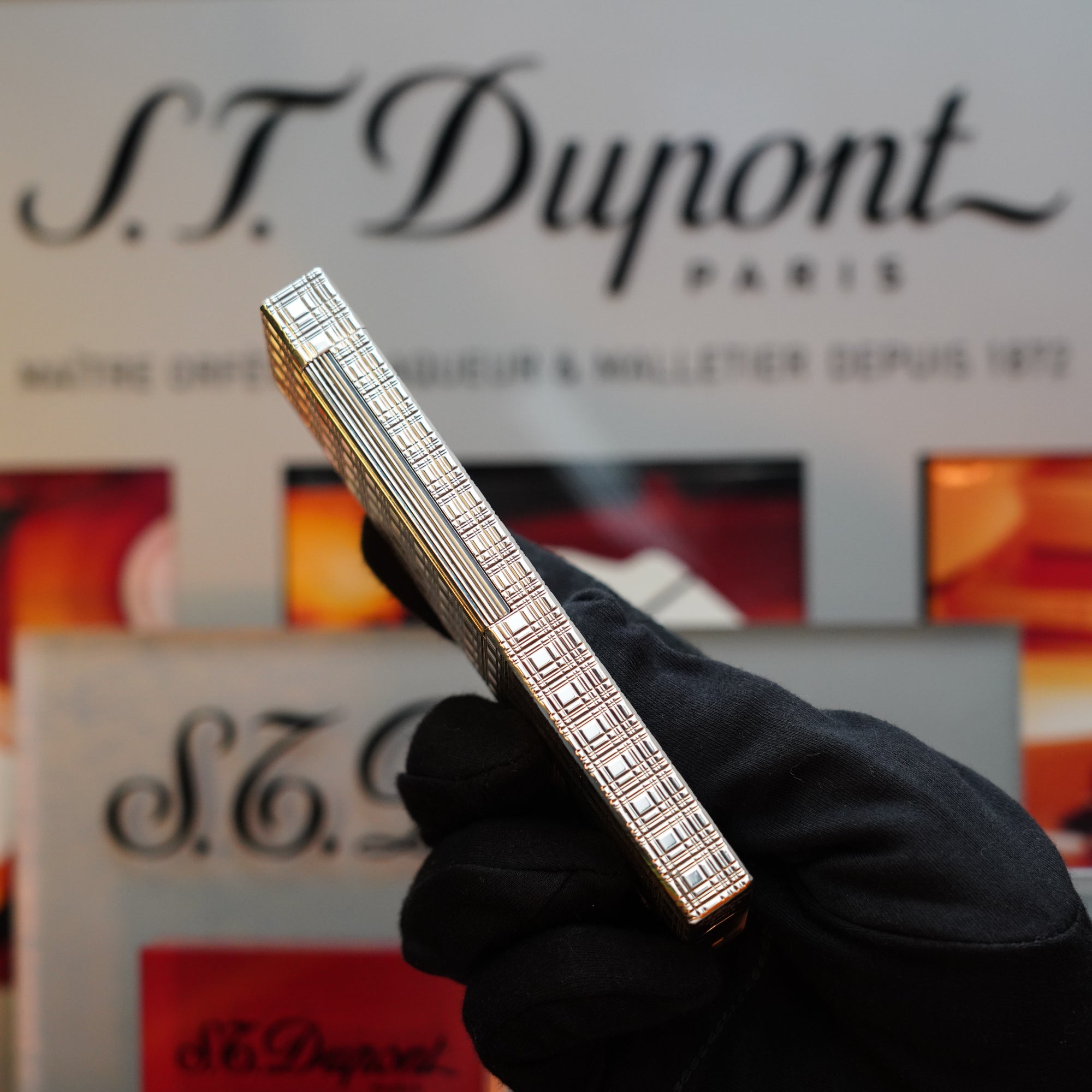 A person wearing black gloves holds a 1960 HERMES Table Lighter with a rare St Dupont Platinum Finish against a blurred background of S.T. Dupont branding materials.