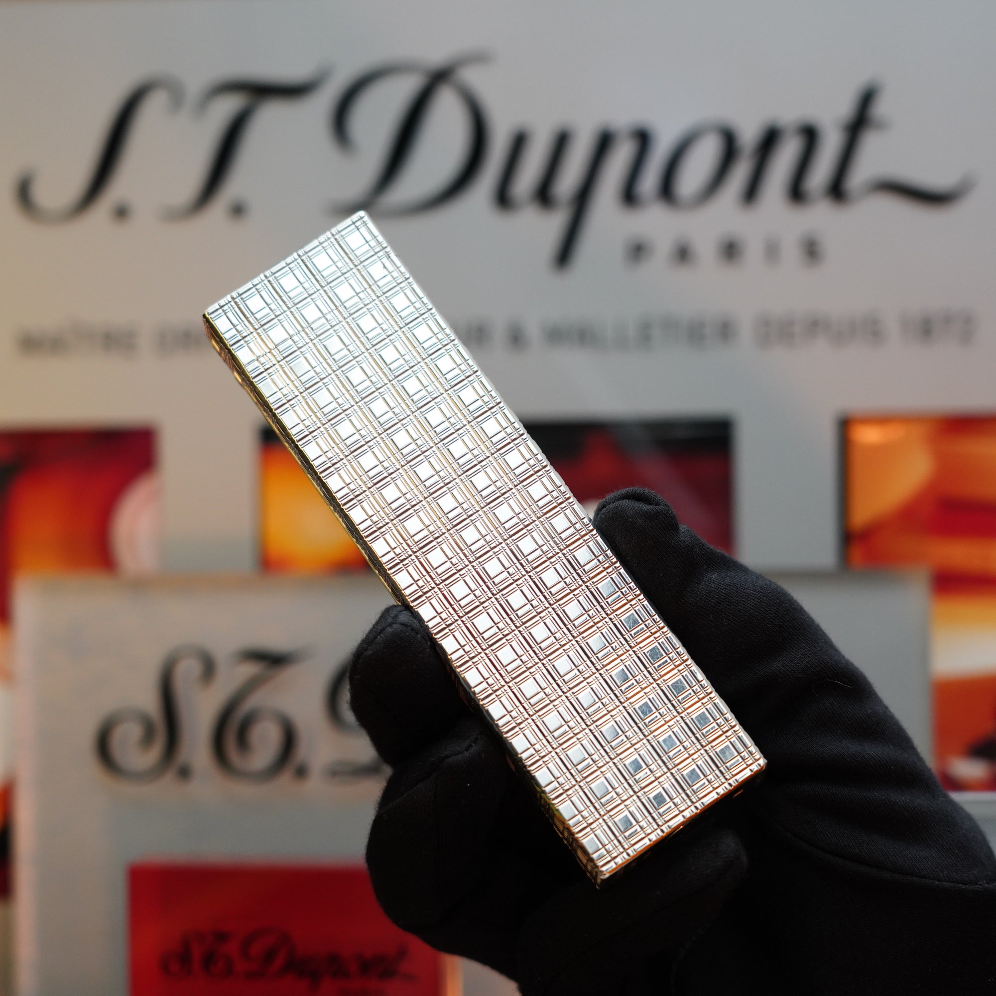 A gloved hand holds a checkered silver lighter in front of an S.T. Dupont Paris sign, evoking the allure of a collector's item with its pristine 1960 HERMES Table Lighter St Dupont Platinum Finish rare pattern.