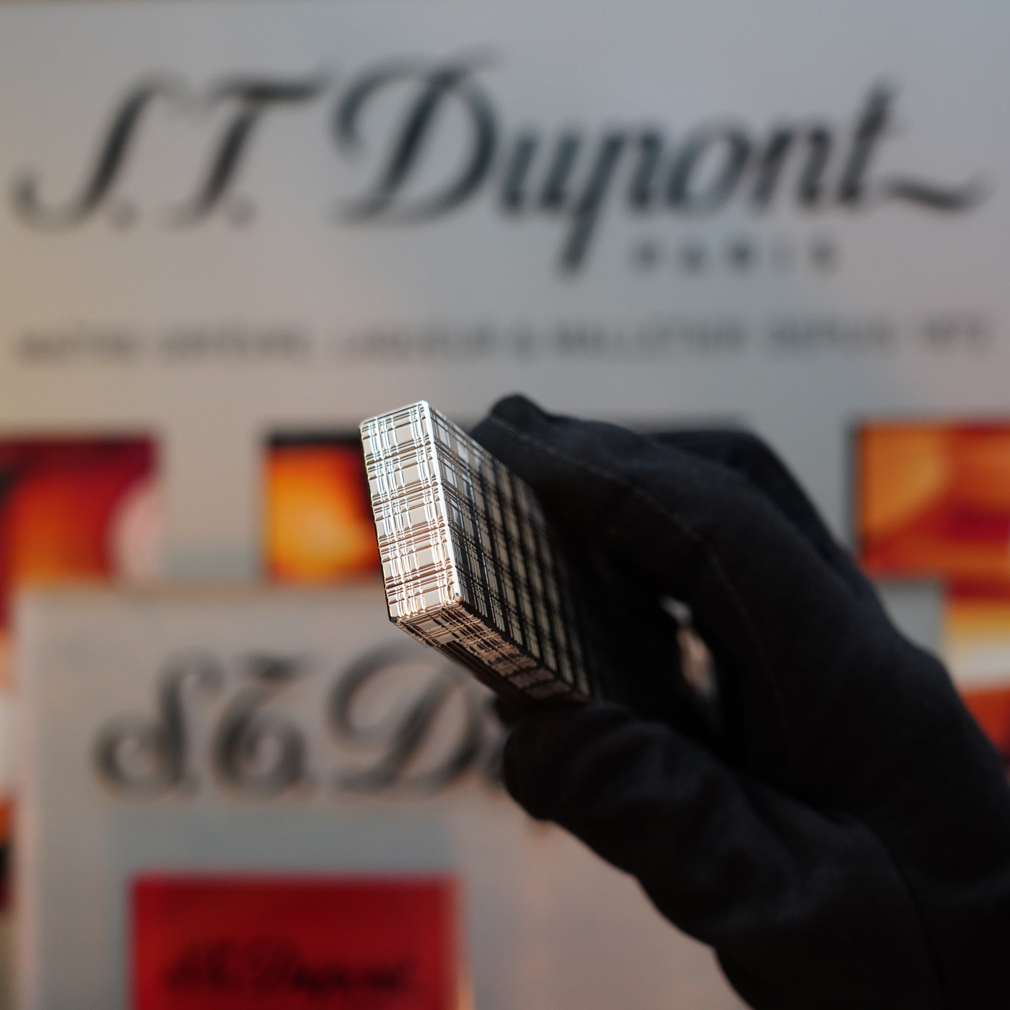 A gloved hand holds a rectangular, patterned object with the 1960 HERMES Table Lighter St Dupont Platinum Finish in front of a blurred background, featuring the text "S.T. Dupont". This elegant piece is truly a collector's item.