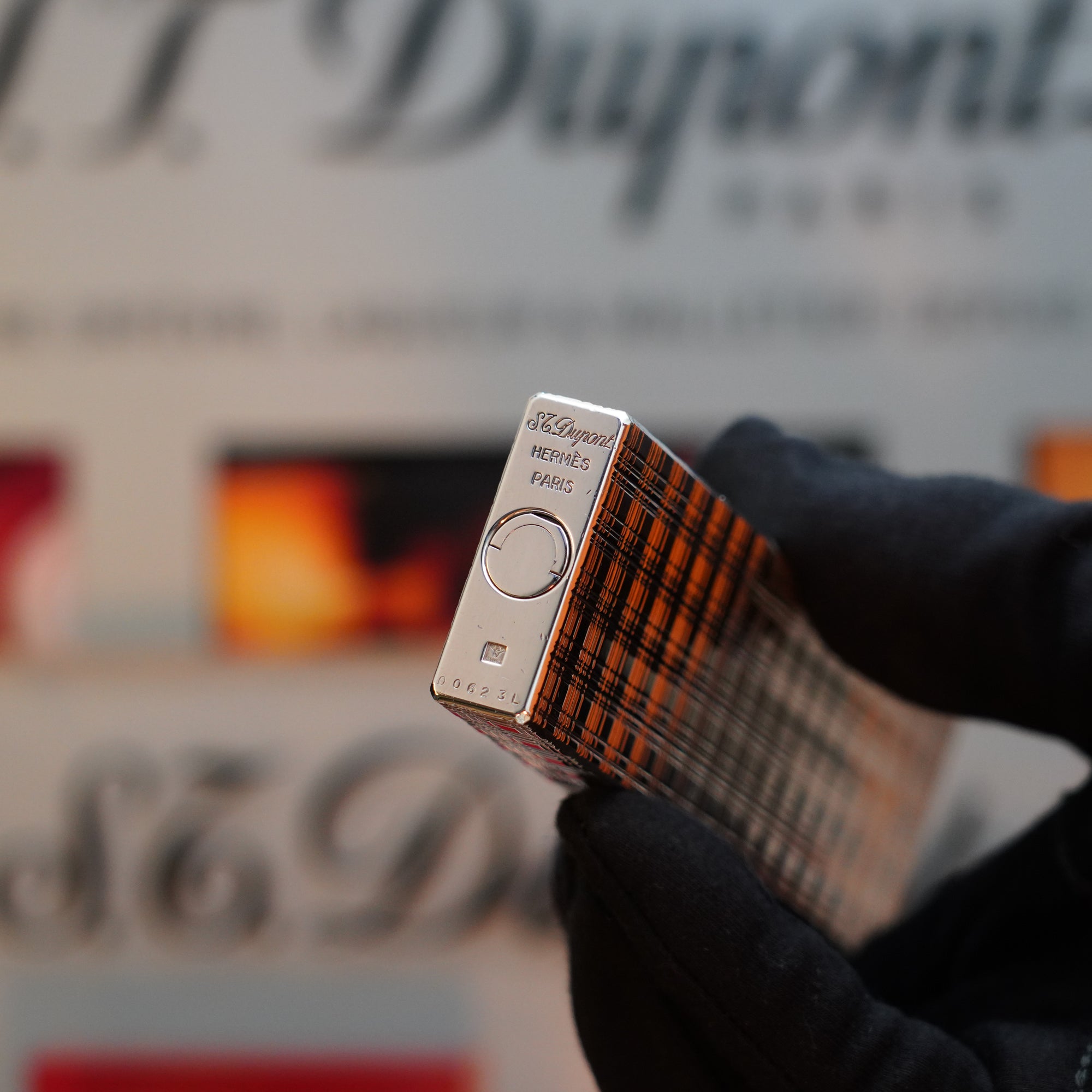 A gloved hand holds a metallic 1960 HERMES Table Lighter St Dupont with a rare plaid pattern, its platinum finish gleaming. The background is blurred and features the S.T. Dupont logo, emphasizing this collector's item for enthusiasts.