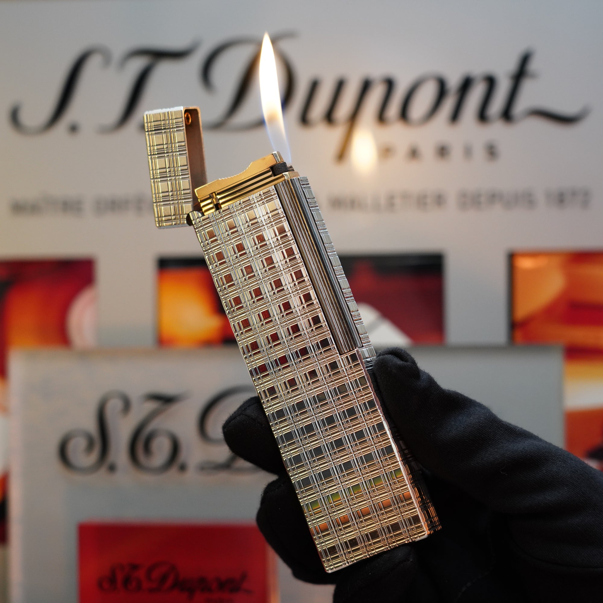 A gloved hand holds a lit silver 1960 HERMES Table Lighter from S.T. Dupont, featuring its rare checkered pattern and exquisite Platinum Finish. The S.T. Dupont branding is visible in the background, highlighting this collector's item.