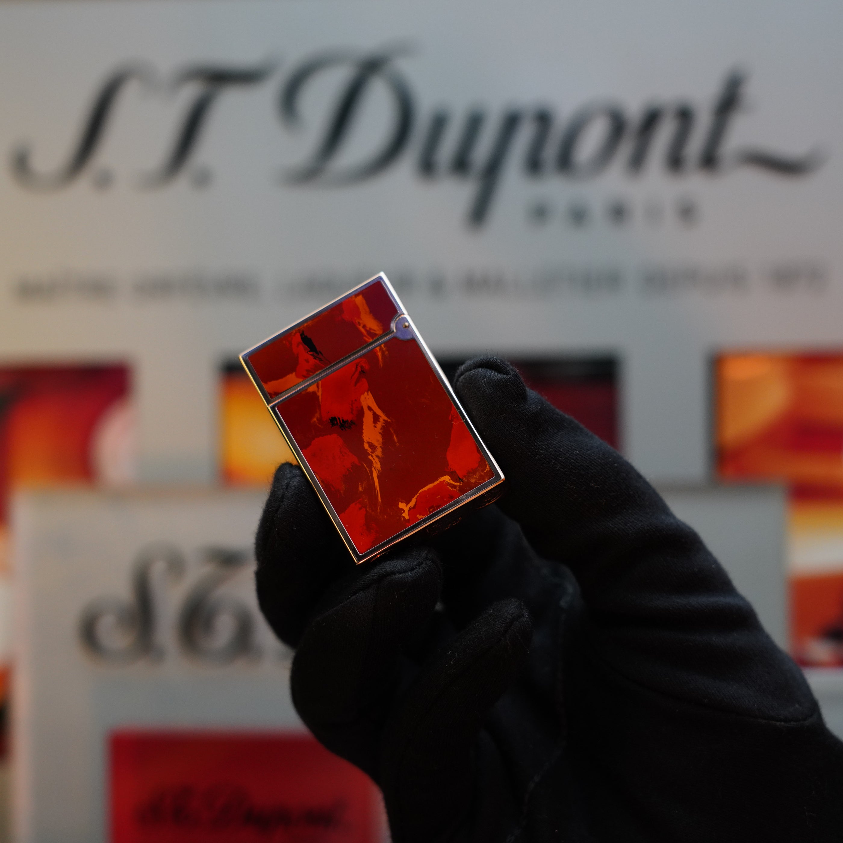 A gloved hand holds a red 1970 S.T. Dupont Picaso 24k Rose Gold Ligne 2 Natural Lacquer rare Ligne 2 Lighter against a blurred background displaying the S.T. Dupont logo, showcasing this exquisite collector's item.