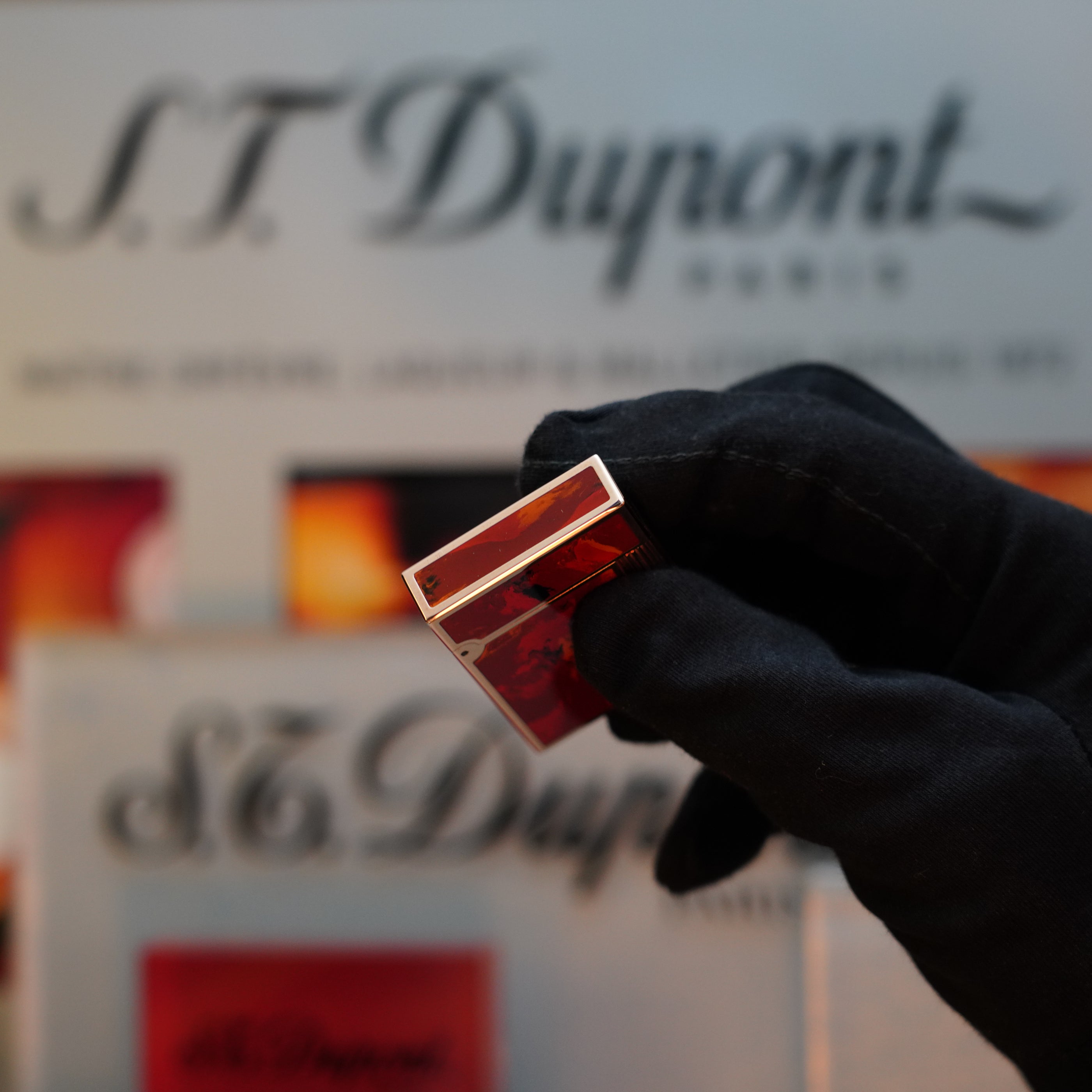 A gloved hand holds a small red and silver object in front of a blurred "S.T. Dupont" sign, revealing a hint of the elegance associated with the 1970 St Dupont Picasso 24k Rose Gold Ligne 2 Natural Lacquer rare ligne 2 Lighter.