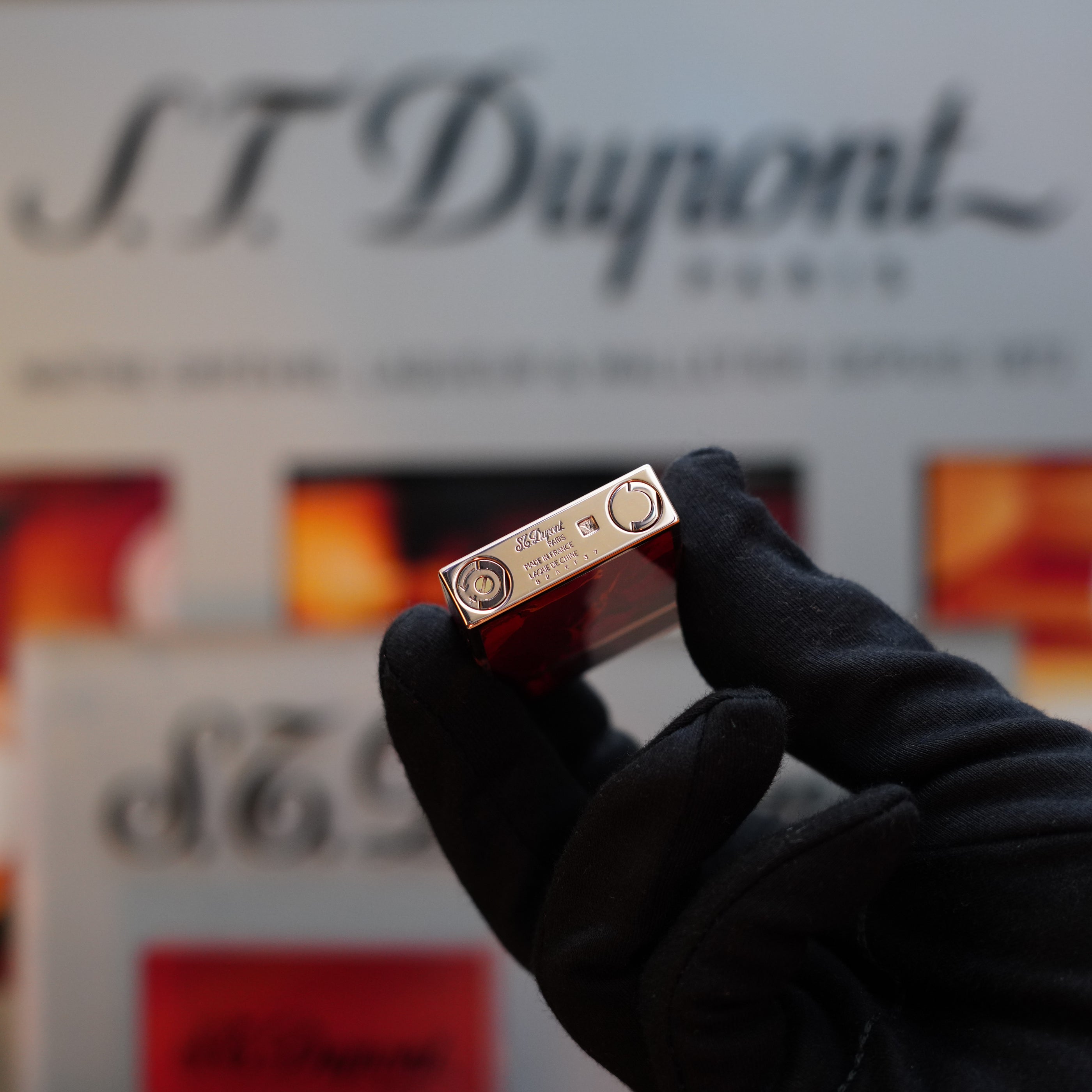 A gloved hand holds a small rectangular object, which appears to be a 1970 St Dupont Picasso 24k Rose Gold Ligne 2 Natural Lacquer rare Ligne 2 lighter, in front of a blurred background with the "S.T. Dupont" logo—an exquisite collector's item.