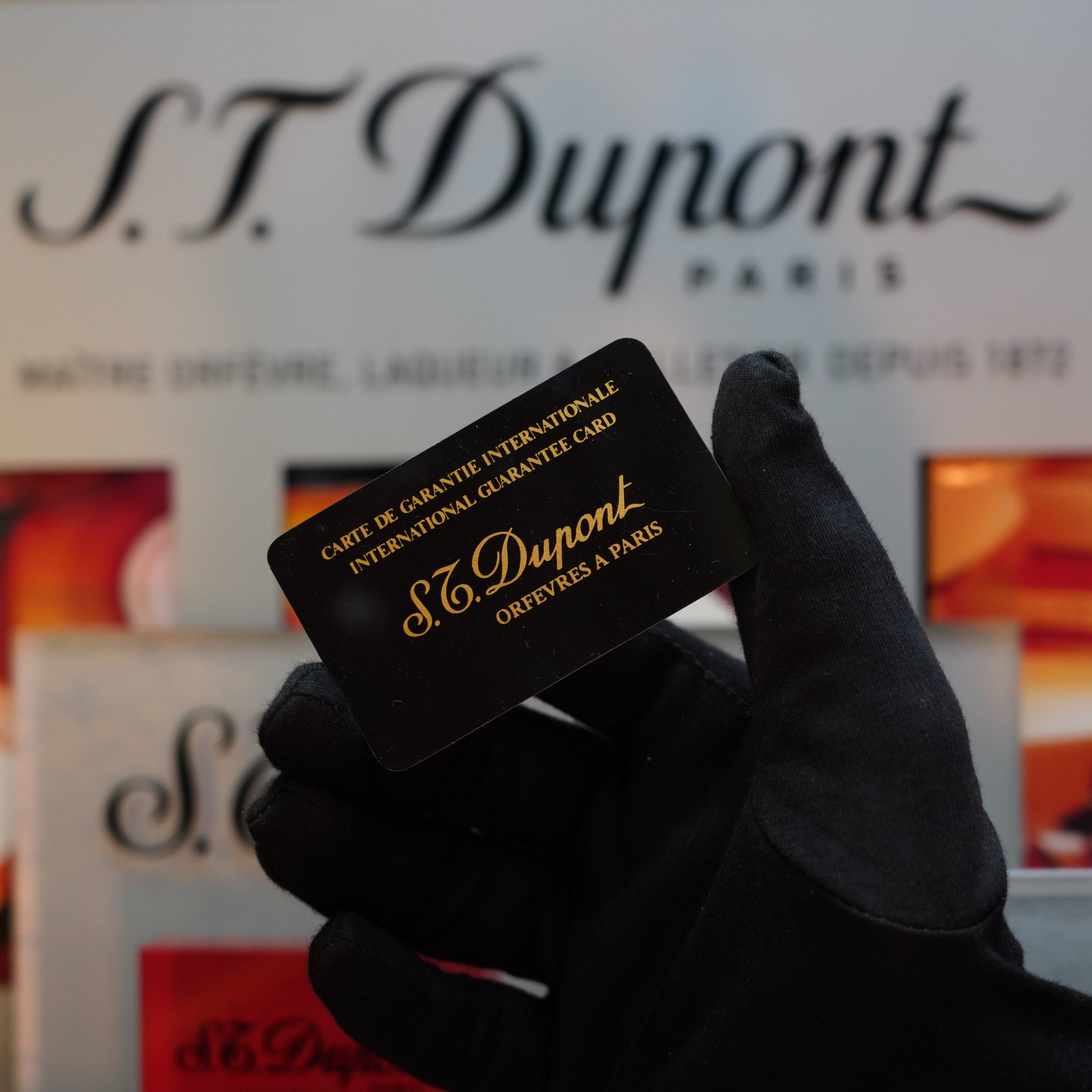 A gloved hand holds an S.T. Dupont international guarantee card against a blurred S.T. Dupont backdrop, showcasing a Collector's Item such as the Rare Vintage 1970 St Dupont Picasso 24k Rose Gold Ligne 2 Natural Lacquer Lighter.