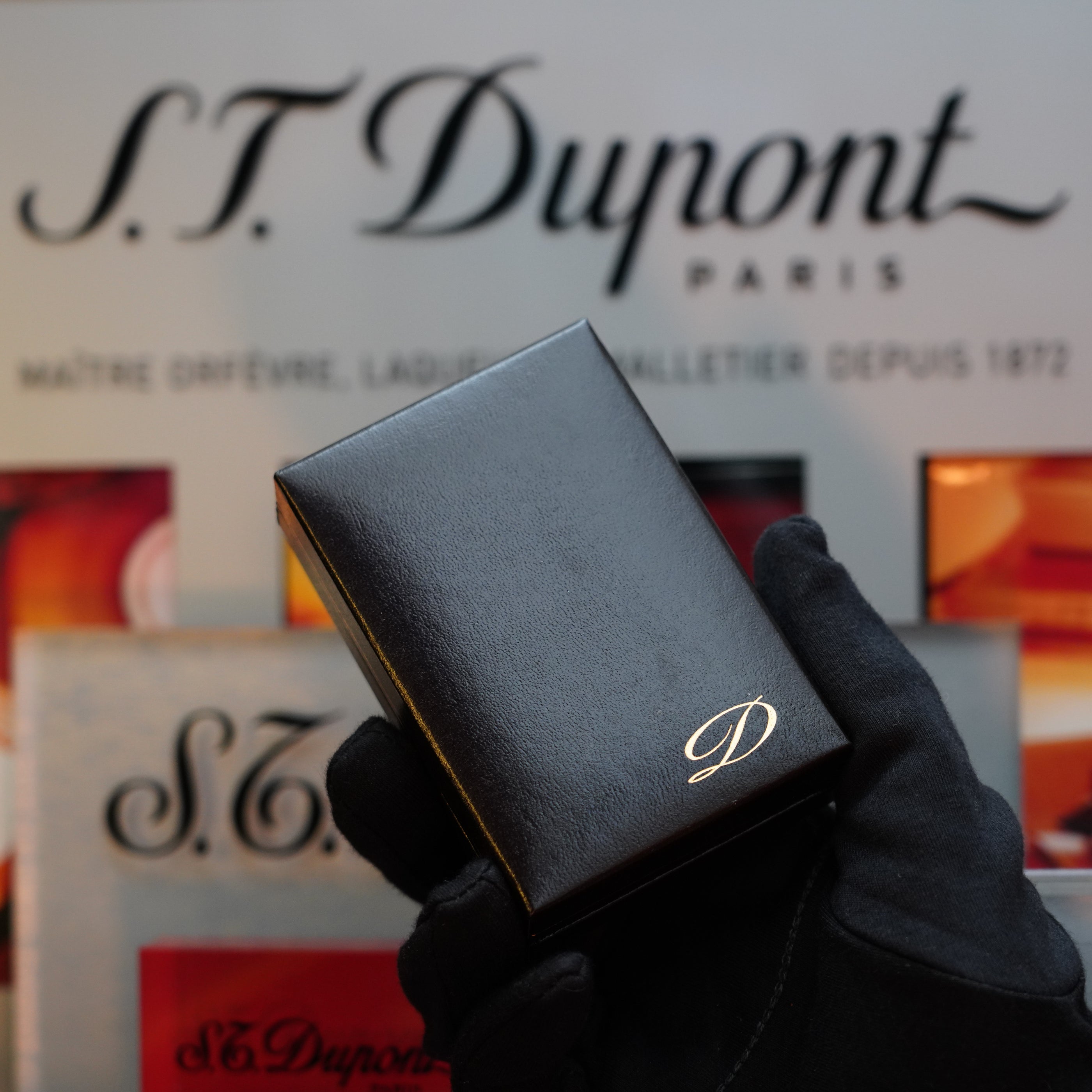 A gloved hand holds a black box adorned with a gold "D" in front of an S.T. Dupont Paris sign—revealing the 1970 St Dupont Picaso 24k Rose Gold Ligne 2 Natural Lacquer lighter, a true collector's item.