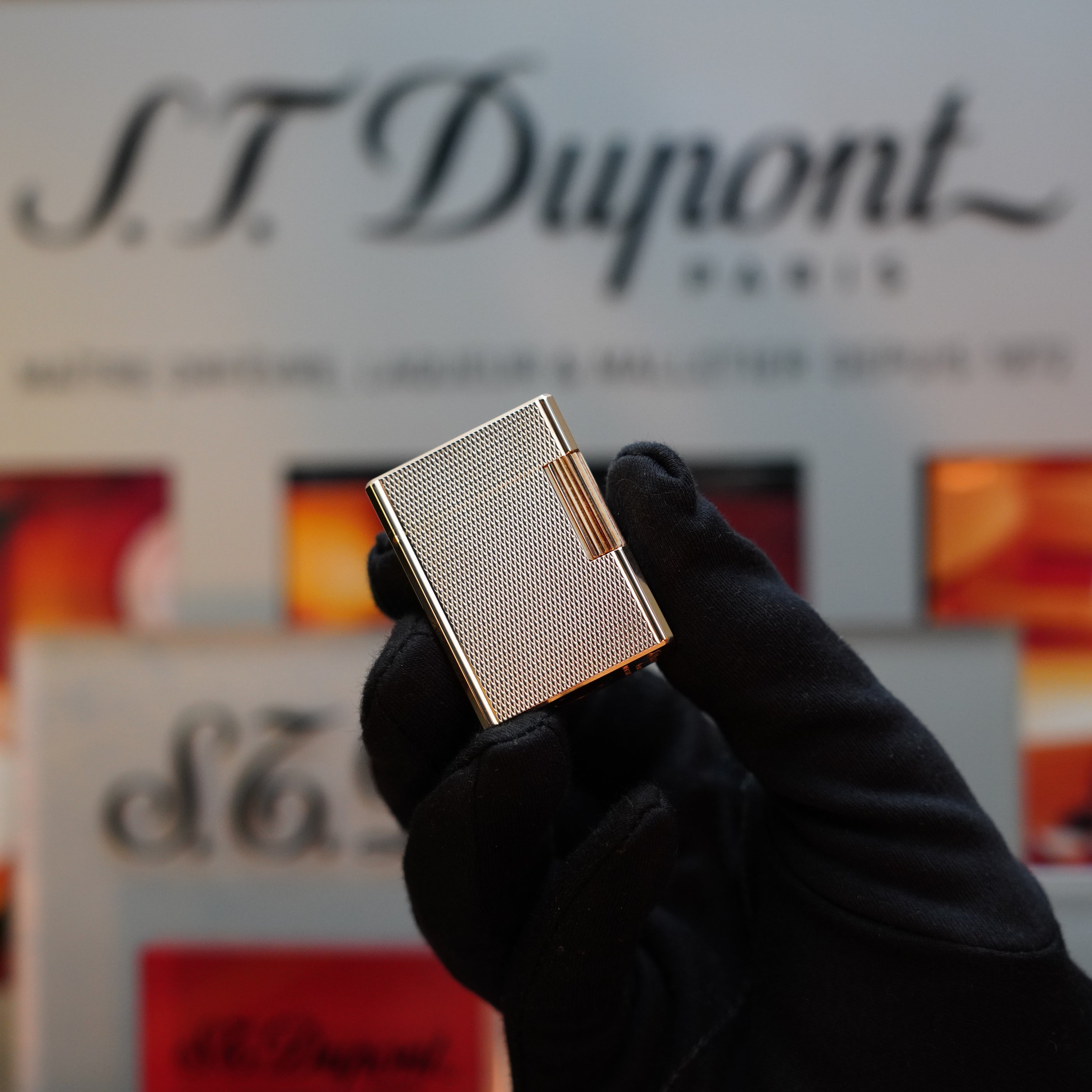 A gloved hand holds a 1970 St Dupont micro Diamond Head Heavy Gold Finish Ligne 1 Lighter. The background displays the S.T. Dupont logo and packaging, showcasing its exquisite craftsmanship.