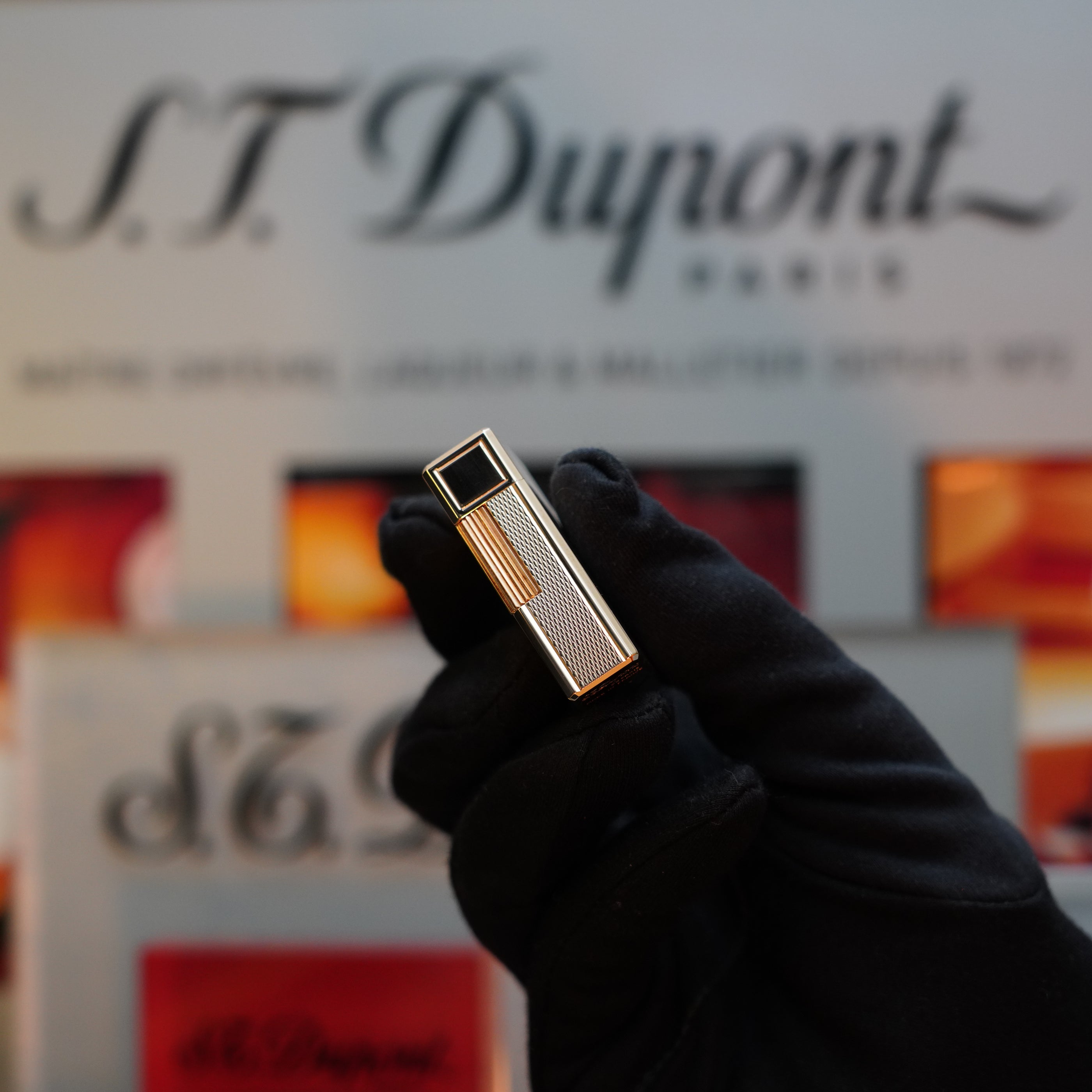 A gloved hand holds a 1970 St Dupont micro Diamond Head Heavy Gold Finish ligne 1 Lighter from S.T. Dupont, set against a blurred logo backdrop.