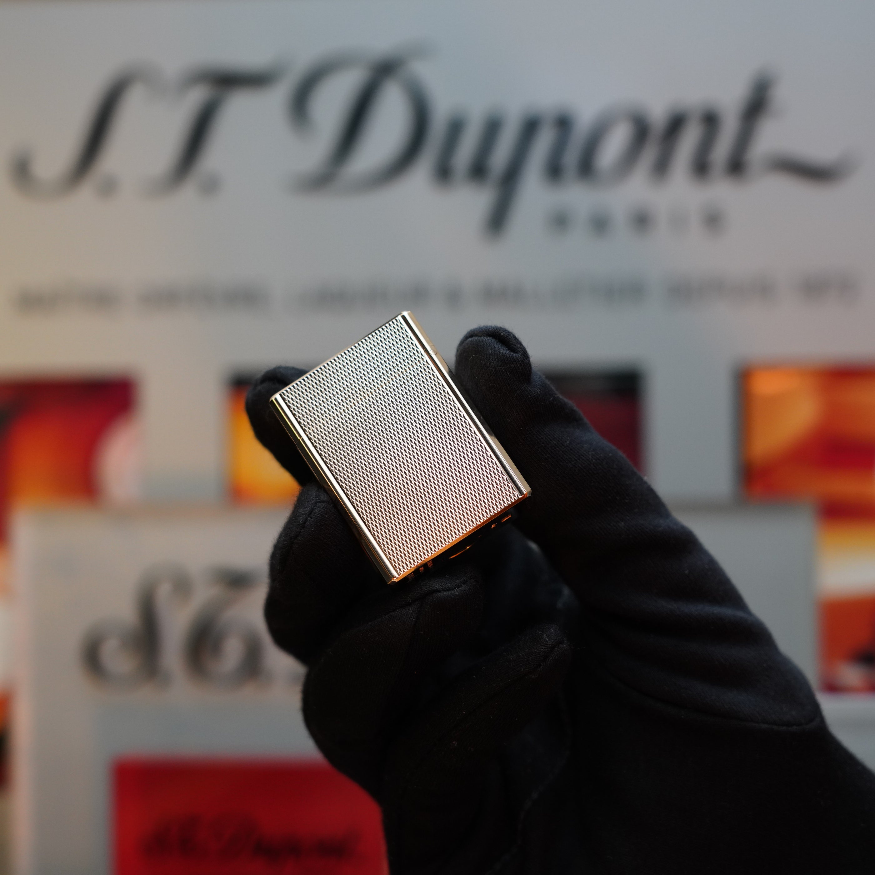 A gloved hand holds a small, rectangular object with a heavy gold finish against a blurred background featuring the branding "S.T. Dupont Paris," reminiscent of the classic 1970 St Dupont micro Diamond Head Heavy Gold Finish Ligne 1 Series lighter.