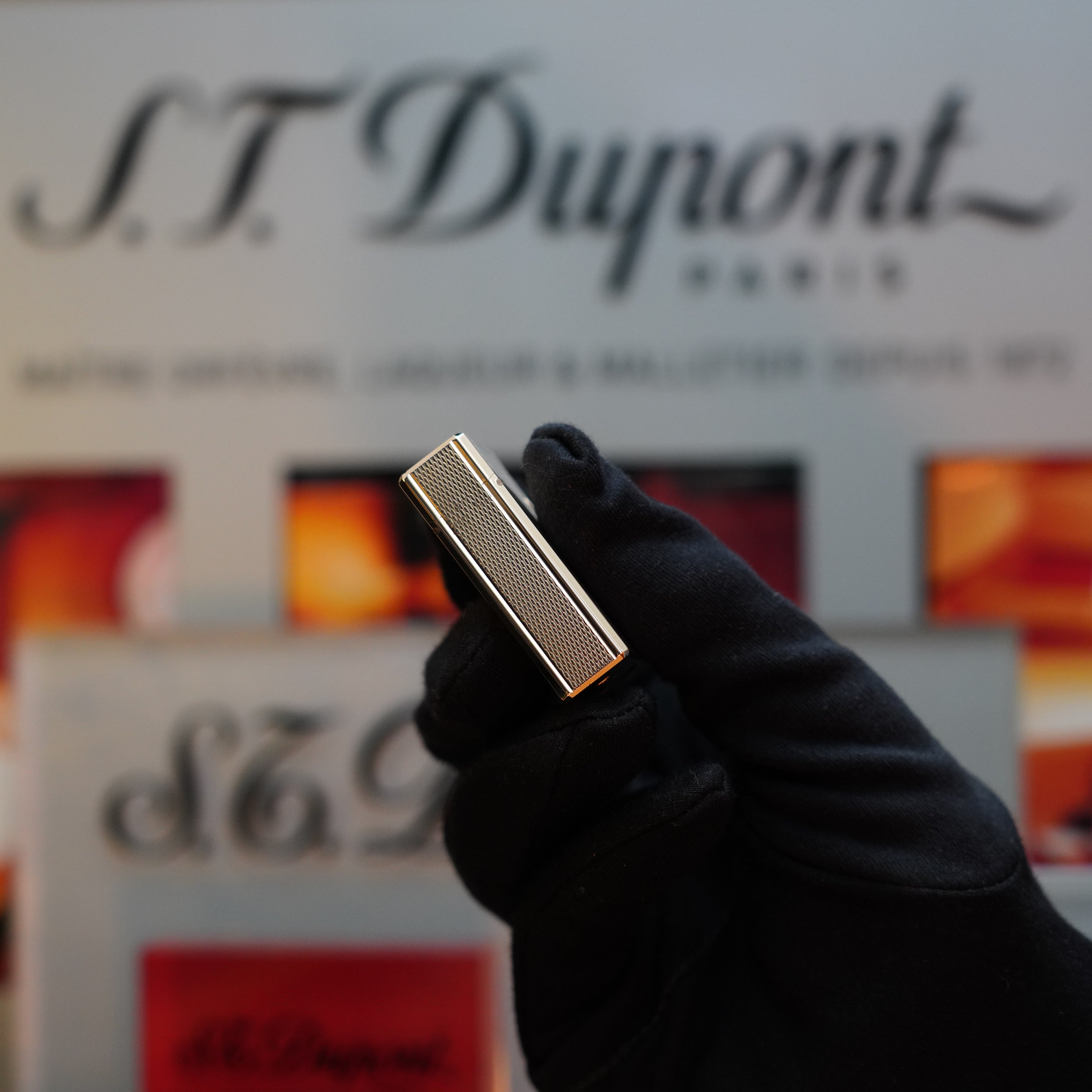 A black-gloved hand holding a heavy gold-finished 1970 S.T. Dupont micro Diamond Head Ligne 1 Lighter in front of a blurred background with S.T. Dupont branding.