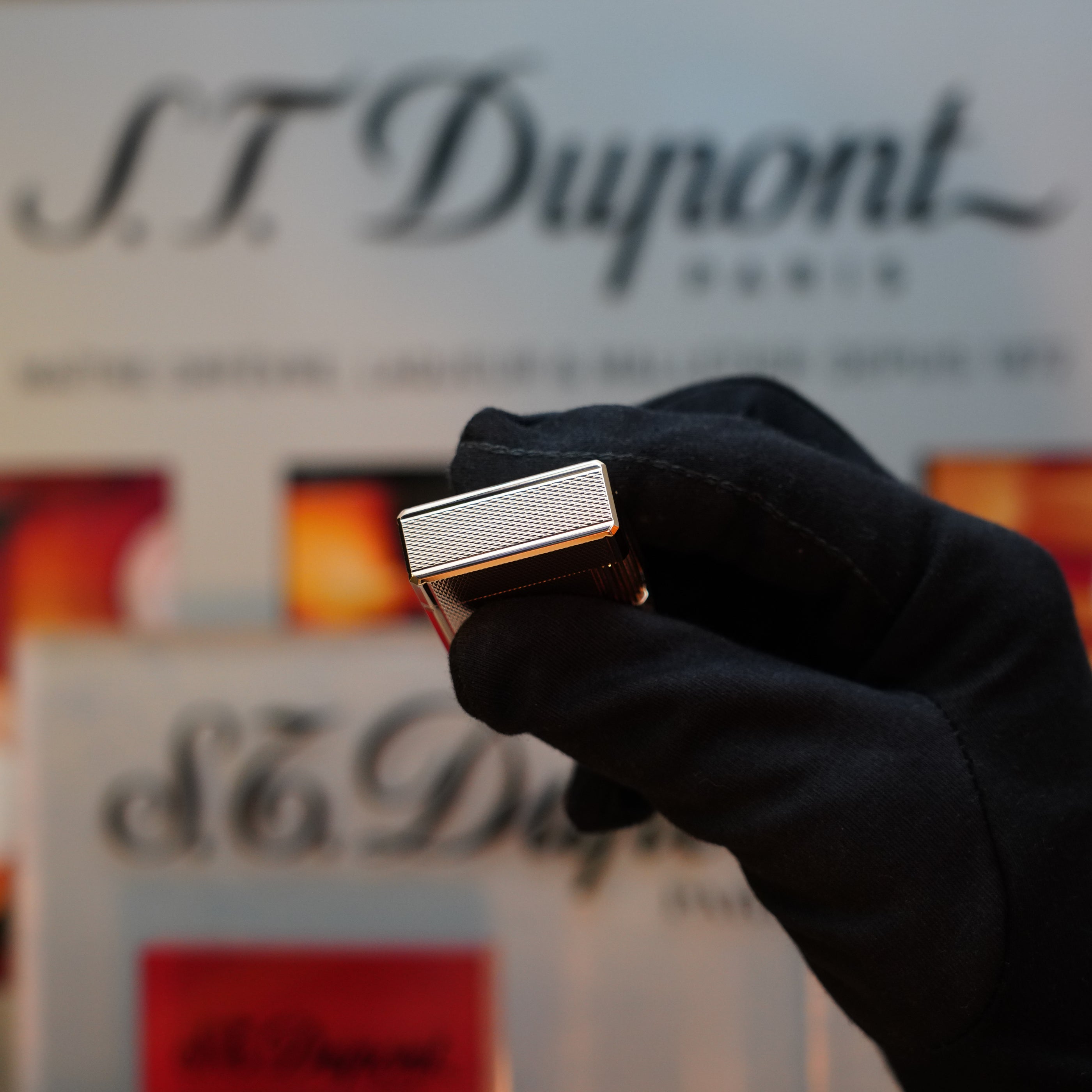 A gloved hand holds a metallic 1970 S.T. Dupont Micro Diamond Head Heavy Gold Finish Ligne 1 Lighter against a blurred background displaying the brand's logo.