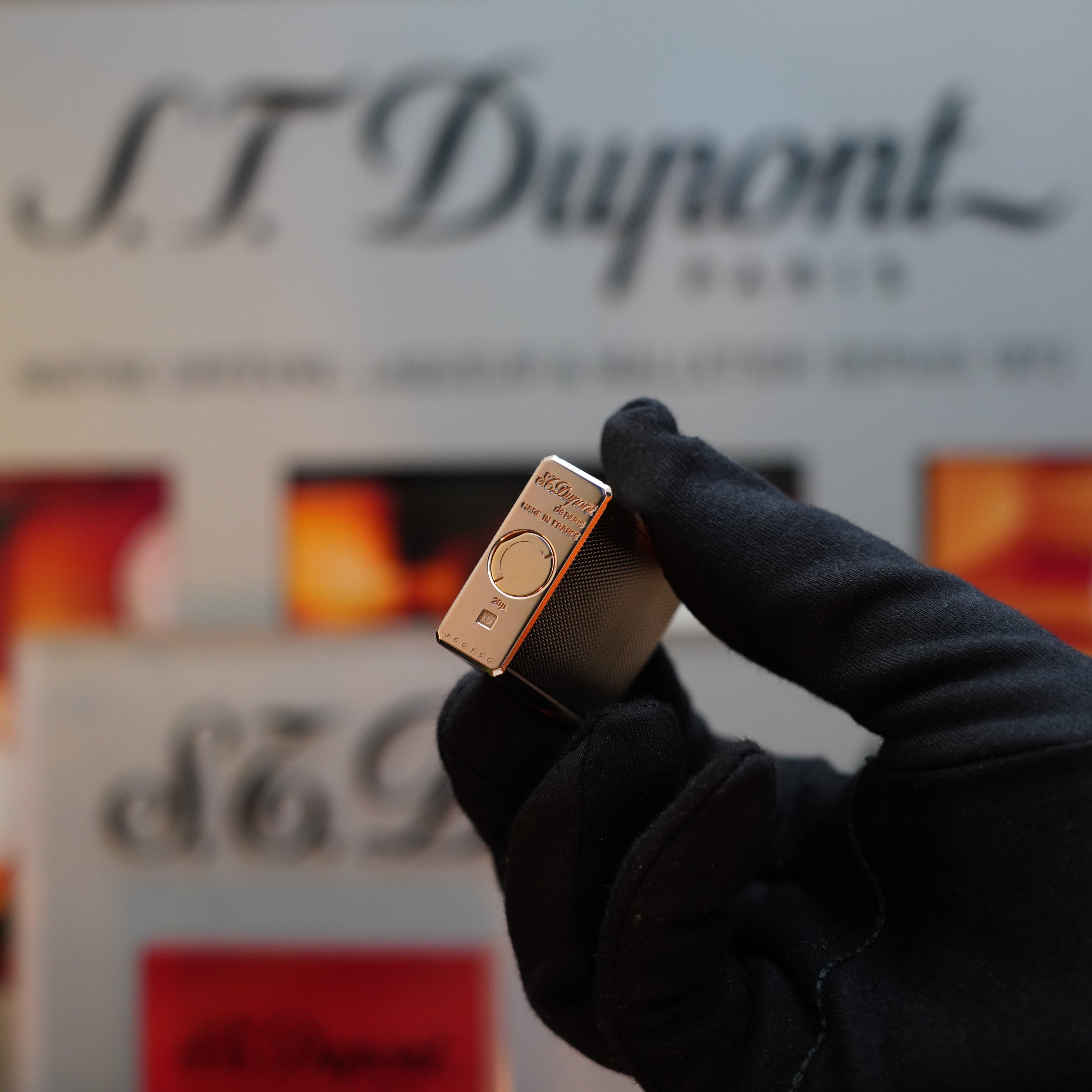 A gloved hand holds a small, rectangular S.T. Dupont product in the foreground, showcasing the elegance of the 1970 St Dupont micro Diamond Head Heavy Gold Finish ligne 1 Lighter, with a blurred S.T. Dupont sign in the background.