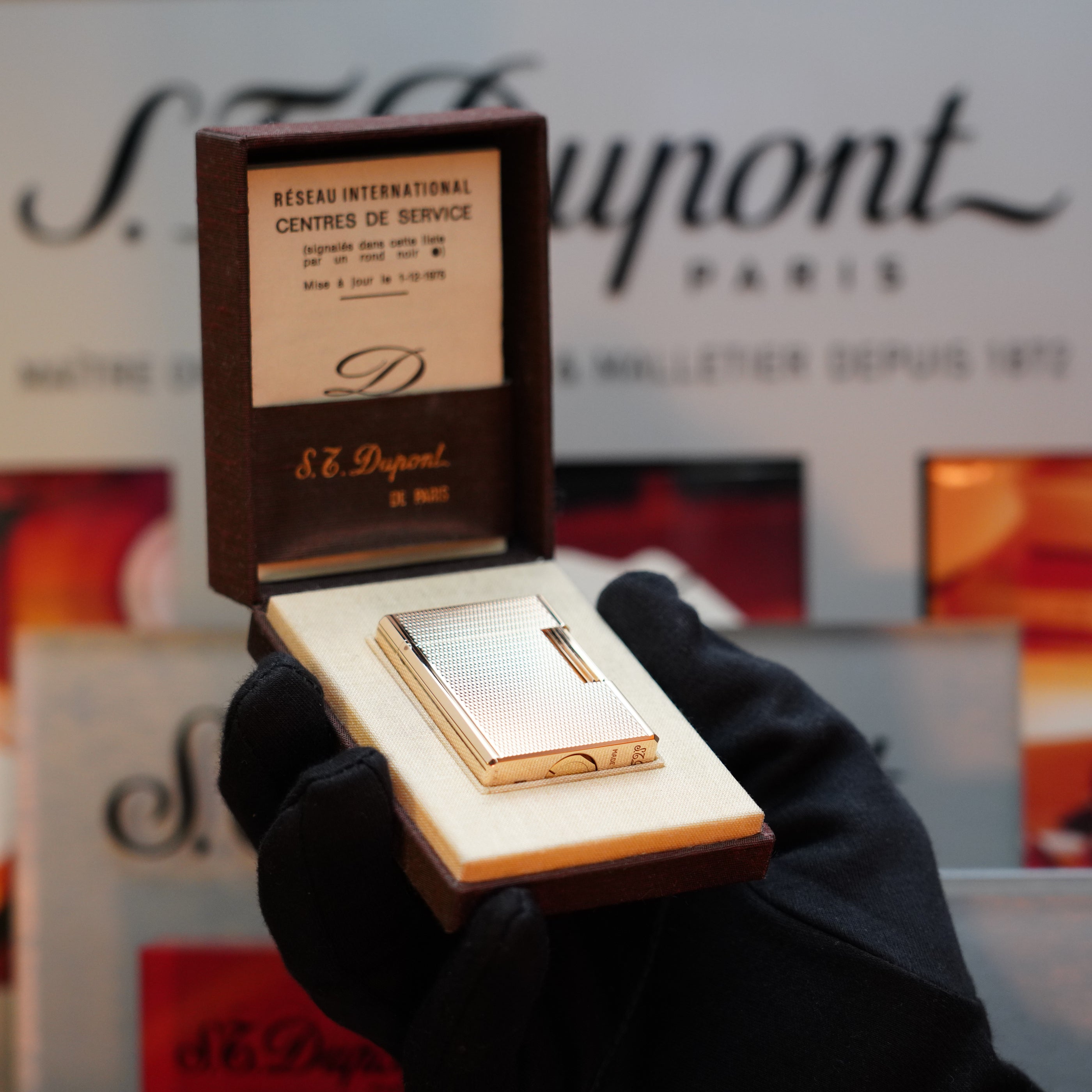 A gloved hand holds an open S.T. Dupont box displaying a 1970 St Dupont micro Diamond Head Heavy Gold Finish ligne 1 Lighter, with the S.T. Dupont branding visible in the background.