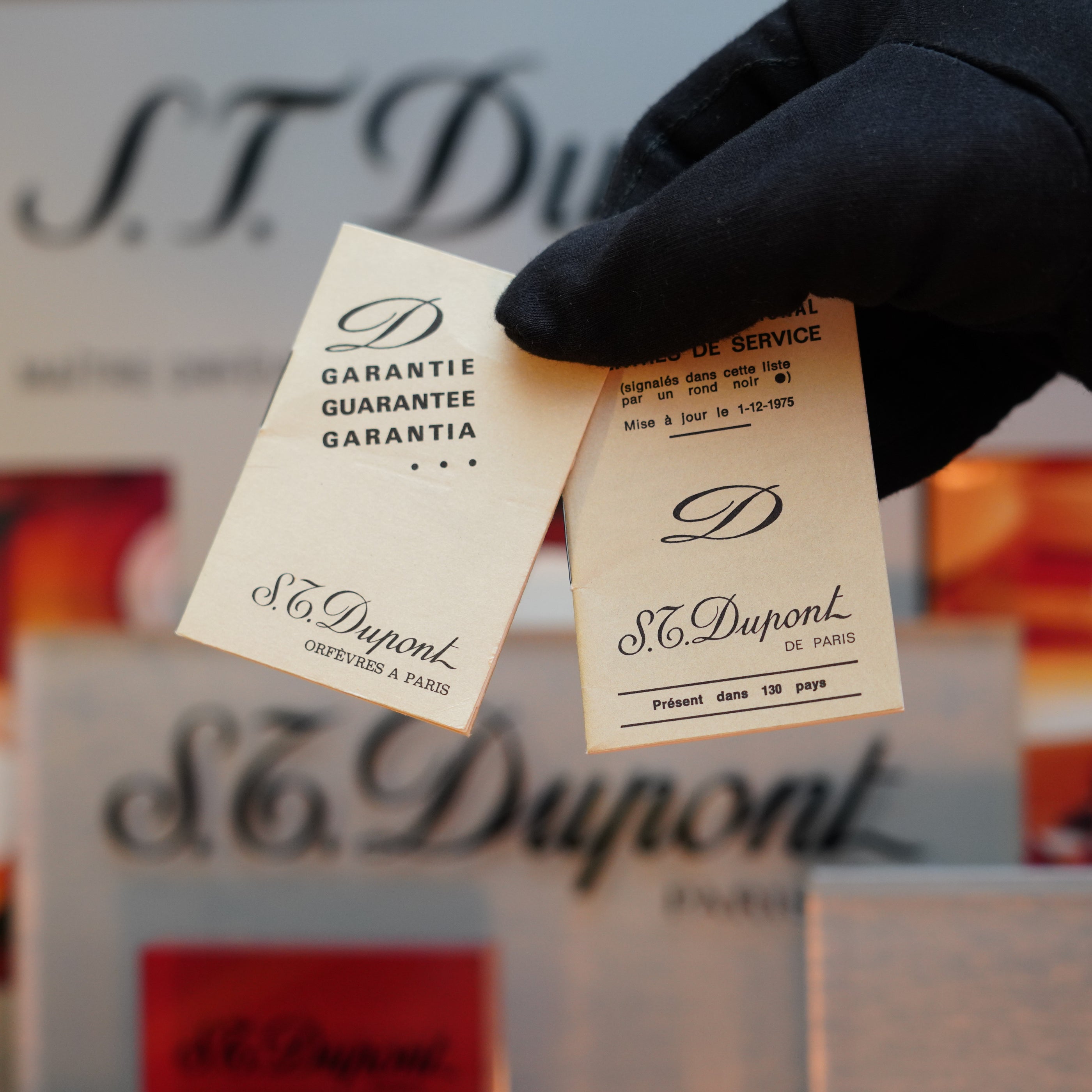 A gloved hand holds two S.T. Dupont warranty cards against a blurred background featuring the S.T. Dupont logo, evoking the elegance of the 1970 St Dupont Micro Diamond Head Heavy Gold Finish Ligne 1 Lighter.