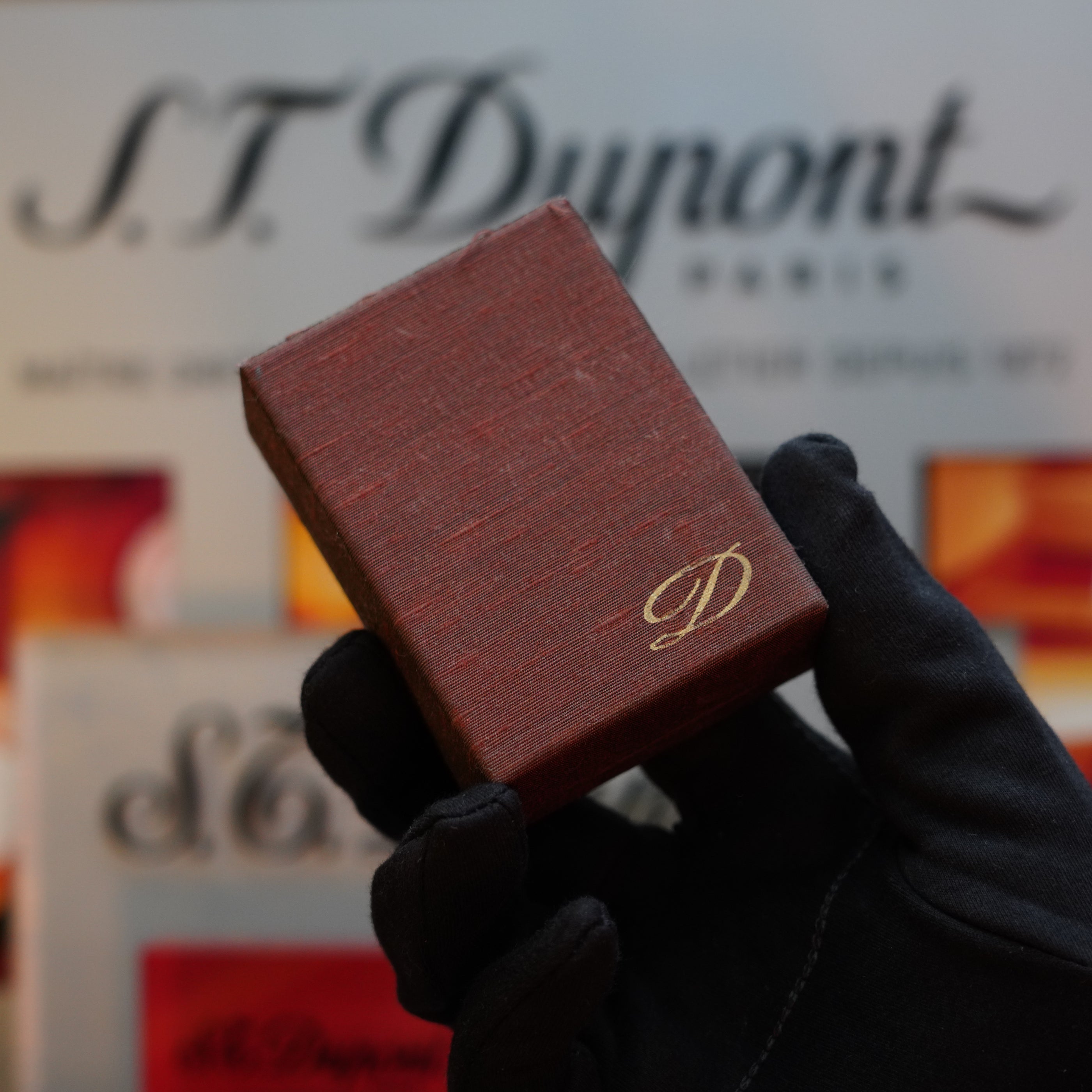 A gloved hand holds a small, brown, textured box with a gold "D" emblem, reminiscent of the 1970 St Dupont micro Diamond Head Heavy Gold Finish ligne 1 Lighter. The background shows a blurred S.T. Dupont Paris logo.